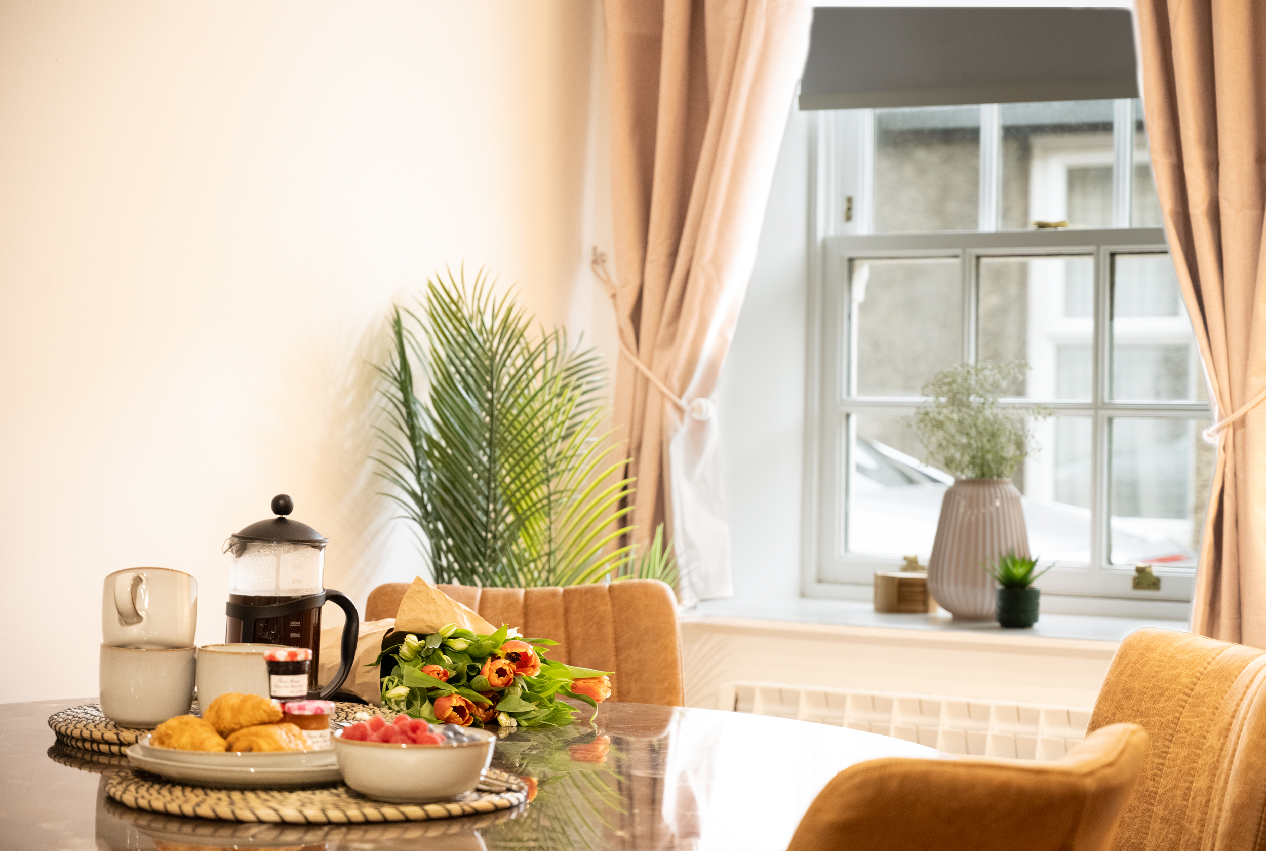 A cozy dining room with a large window, beige curtains, and a plant. A table with breakfast items, flowers, and kitchen appliances is visible.