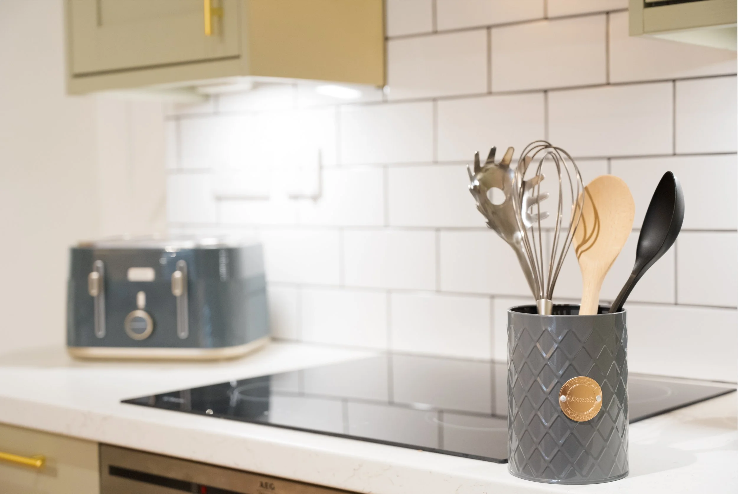 Kitchen countertop with a black cup holding various cooking utensils including a whisk, spatula, and wooden spoon. In the background, a kitchen stove and white tiled backsplash are visible.