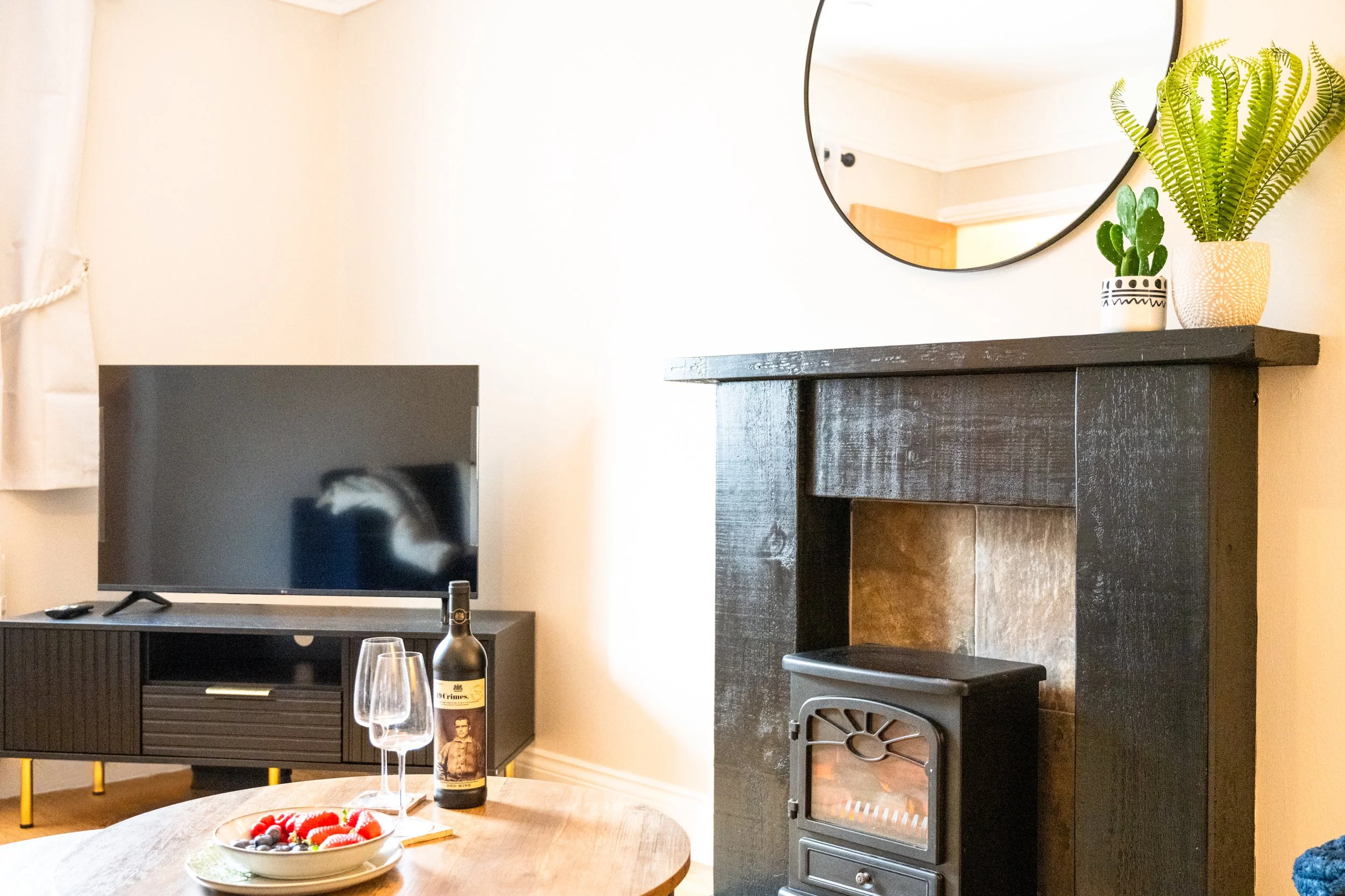 Living room with flatscreen TV on black stand, a round wooden table with a bowl of berries, wine, and glasses, fireplace with black mantel, and wall decor including a mirror and potted plants.