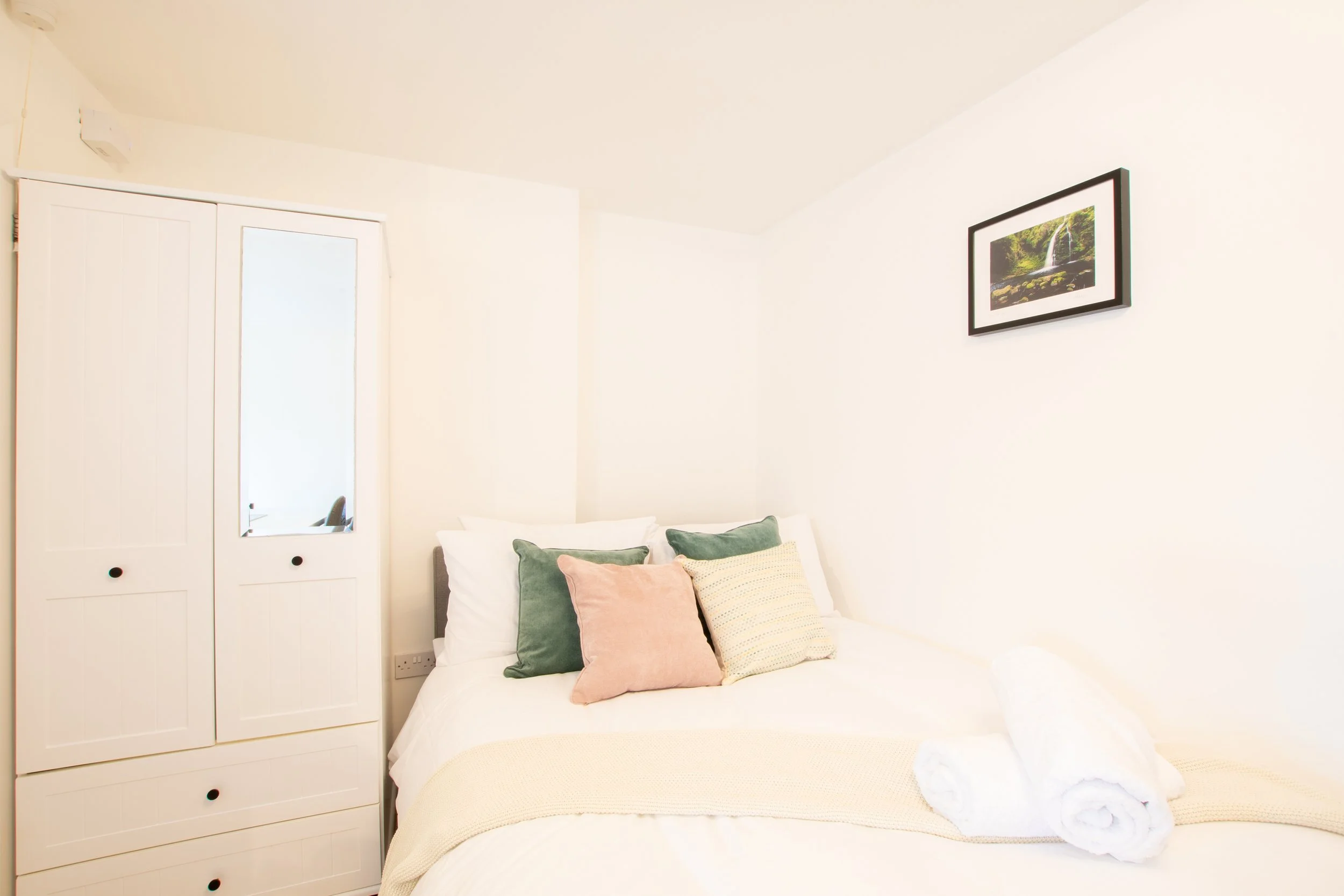 A bedroom with a bed covered in white linens, adorned with pastel-colored pillows including green, pink, and beige. To the left, there is a white closet with a mirror on one of the doors. On the wall above the bed, there is a framed picture of a wate