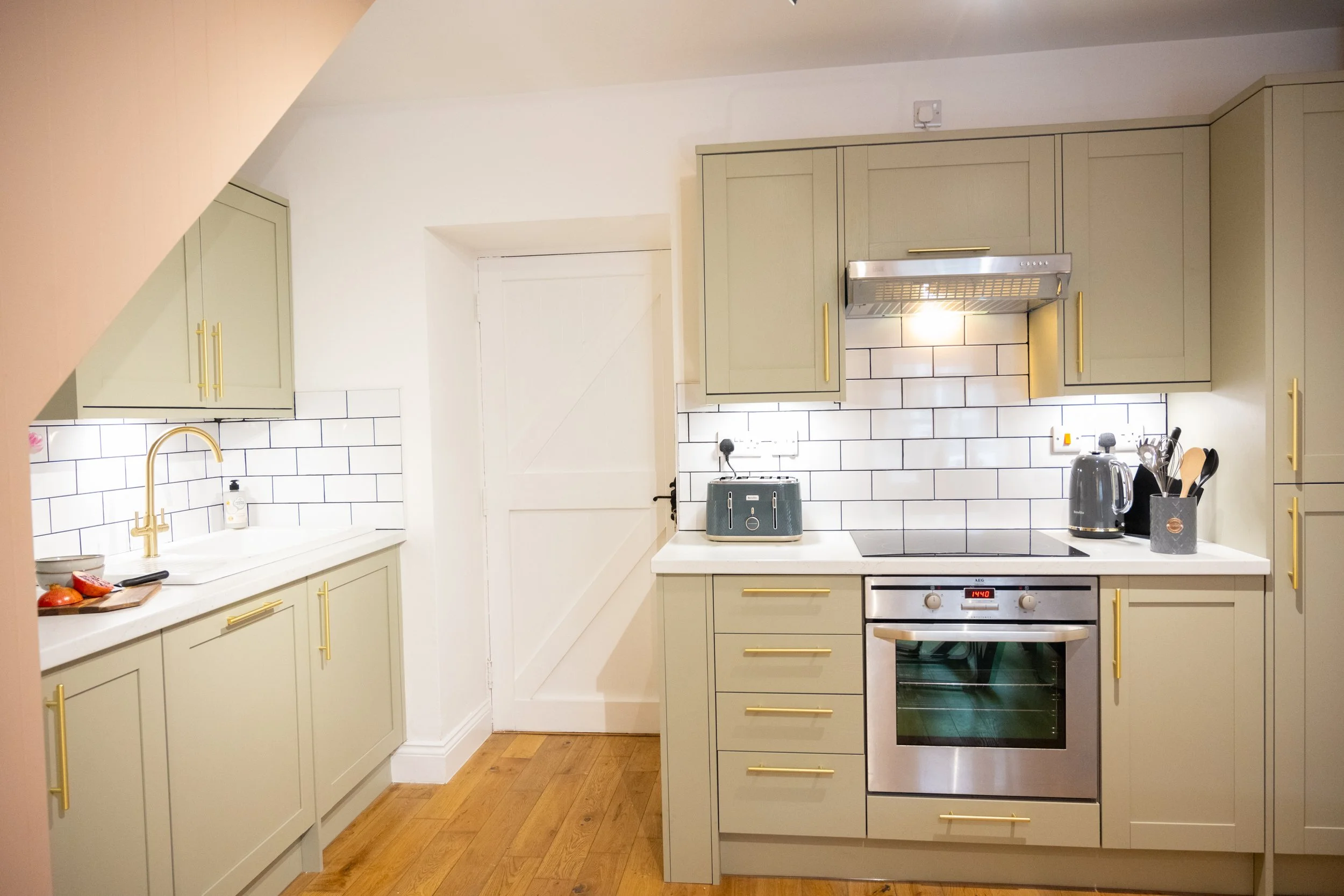 A modern kitchen with light green cabinets with gold handles, white countertops, and a white tiled backsplash. There is a built-in oven, a stovetop, a kettle, and various kitchen utensils. A small window and a door are also visible.