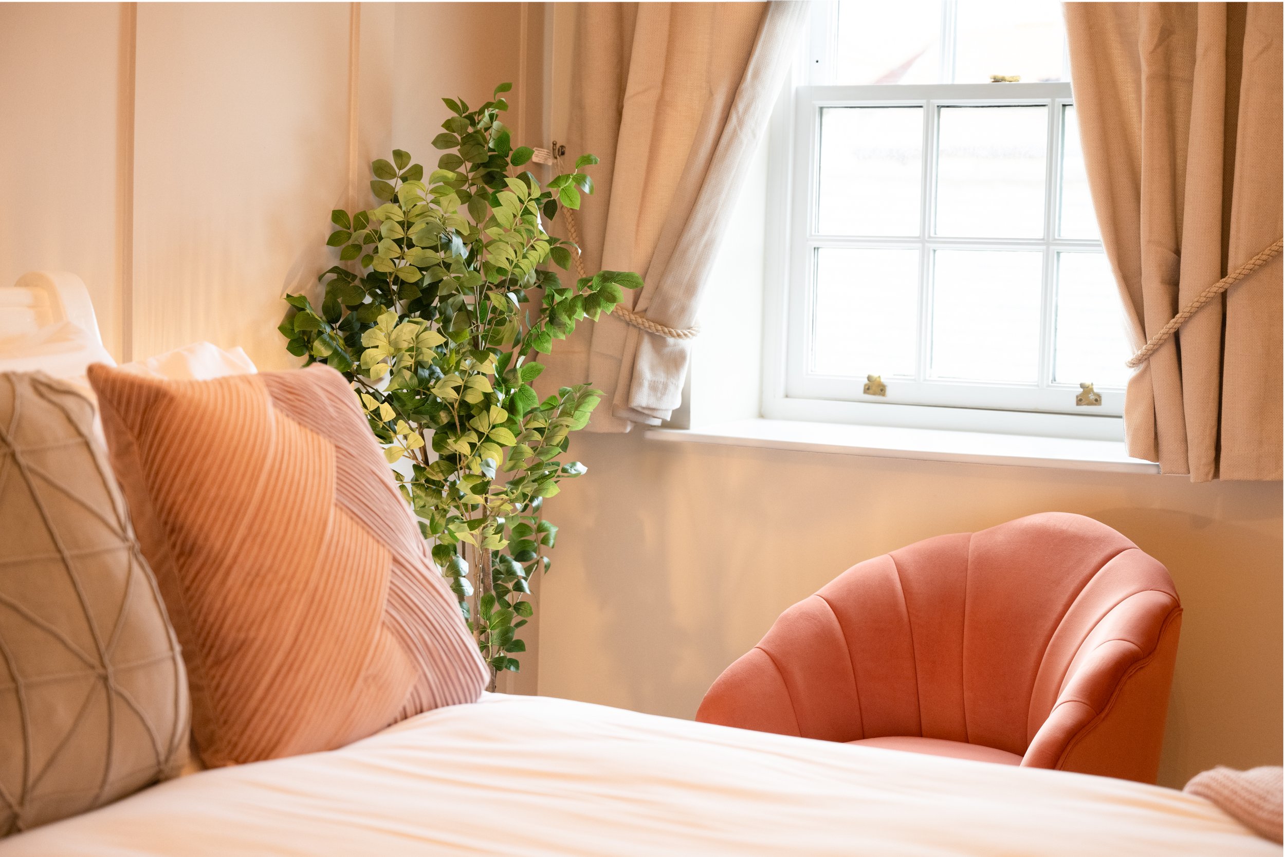 A cozy corner in a room with a large window, beige curtains, a potted plant, a pink upholstered chair, and pillows on a bed or bench.