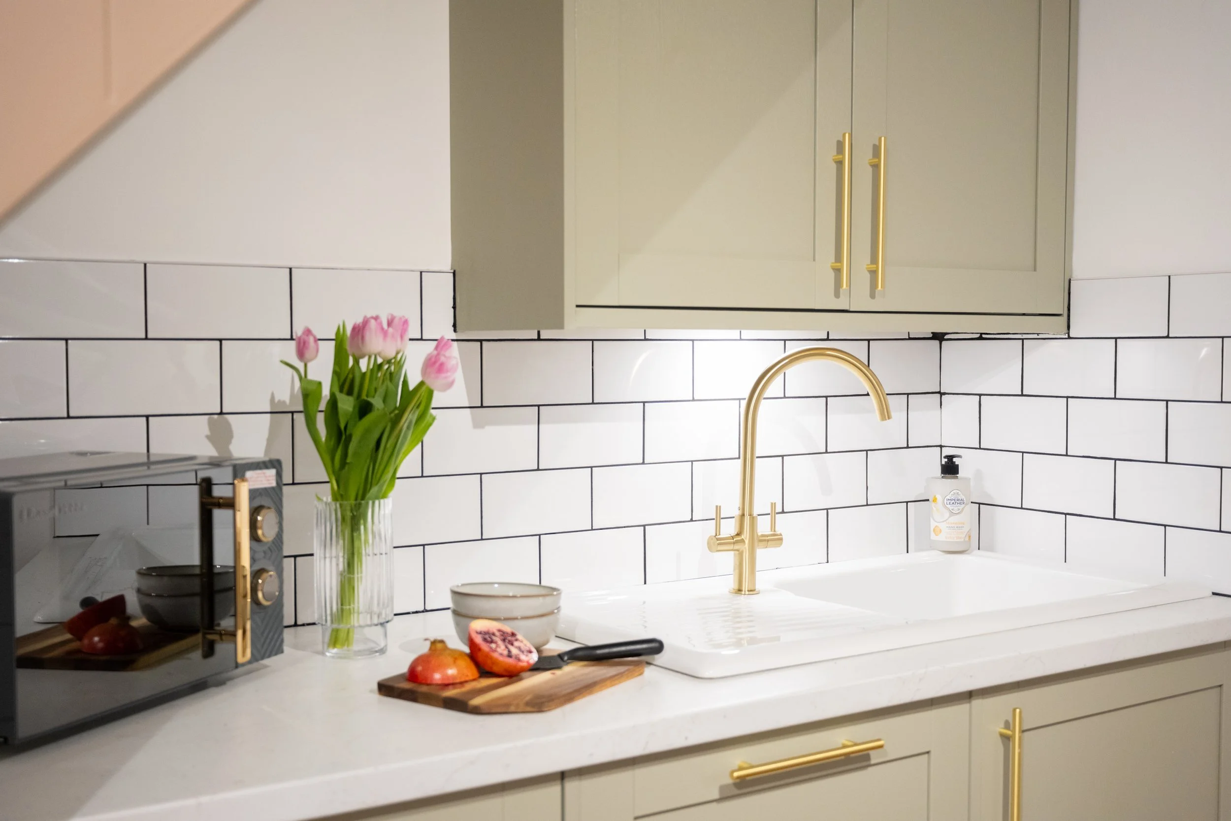 Kitchen with white subway tile backsplash, gold faucet, beige cabinets, a clear vase with pink tulips, a black knife, halved pomegranate, pomegranate seeds, a jar of lotion, and a small bowl on the white countertop.