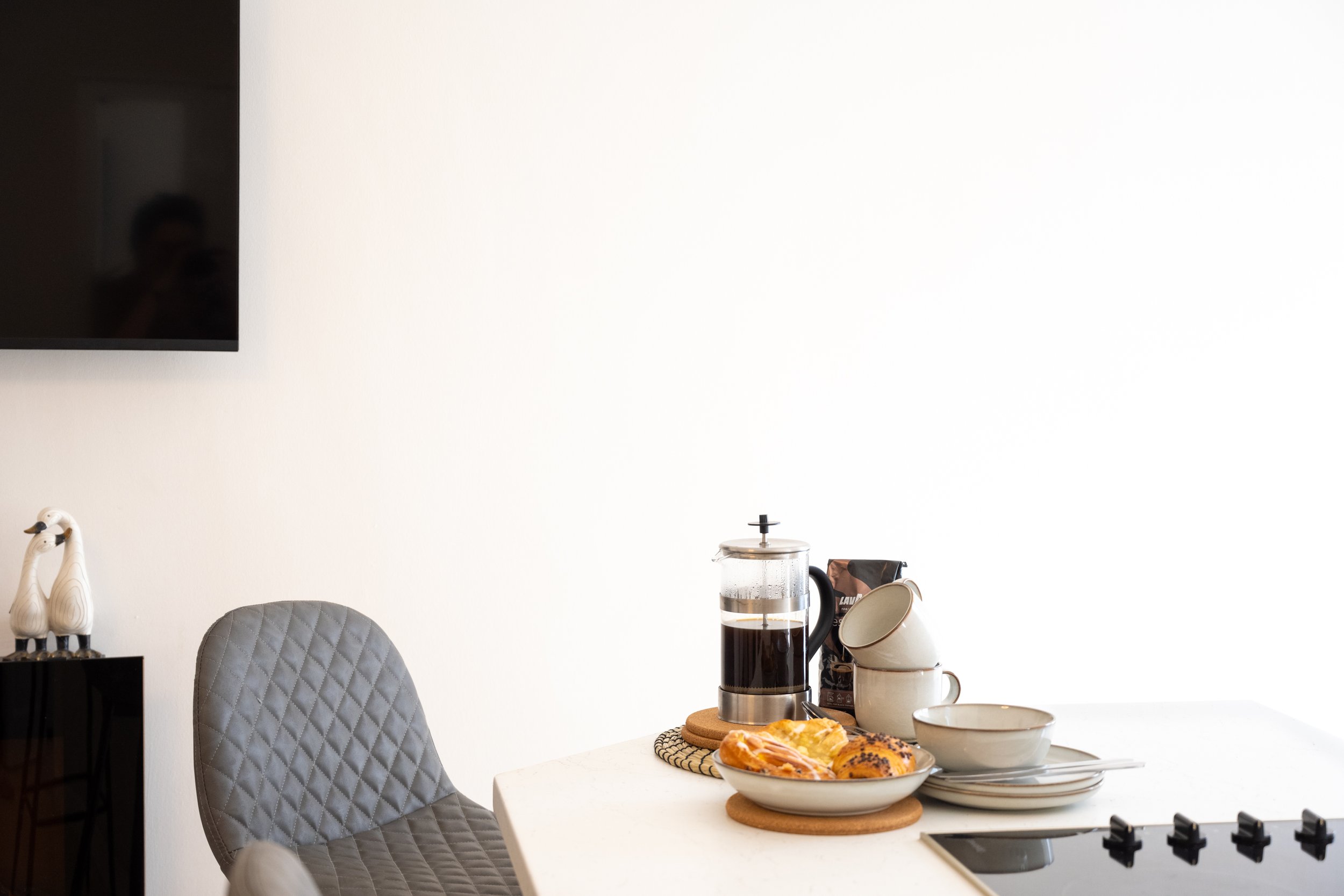 Kitchen table with glass French press coffee maker, plate of pastries, and cups next to a coffee bag; part of a chair and wall-mounted TV are visible.