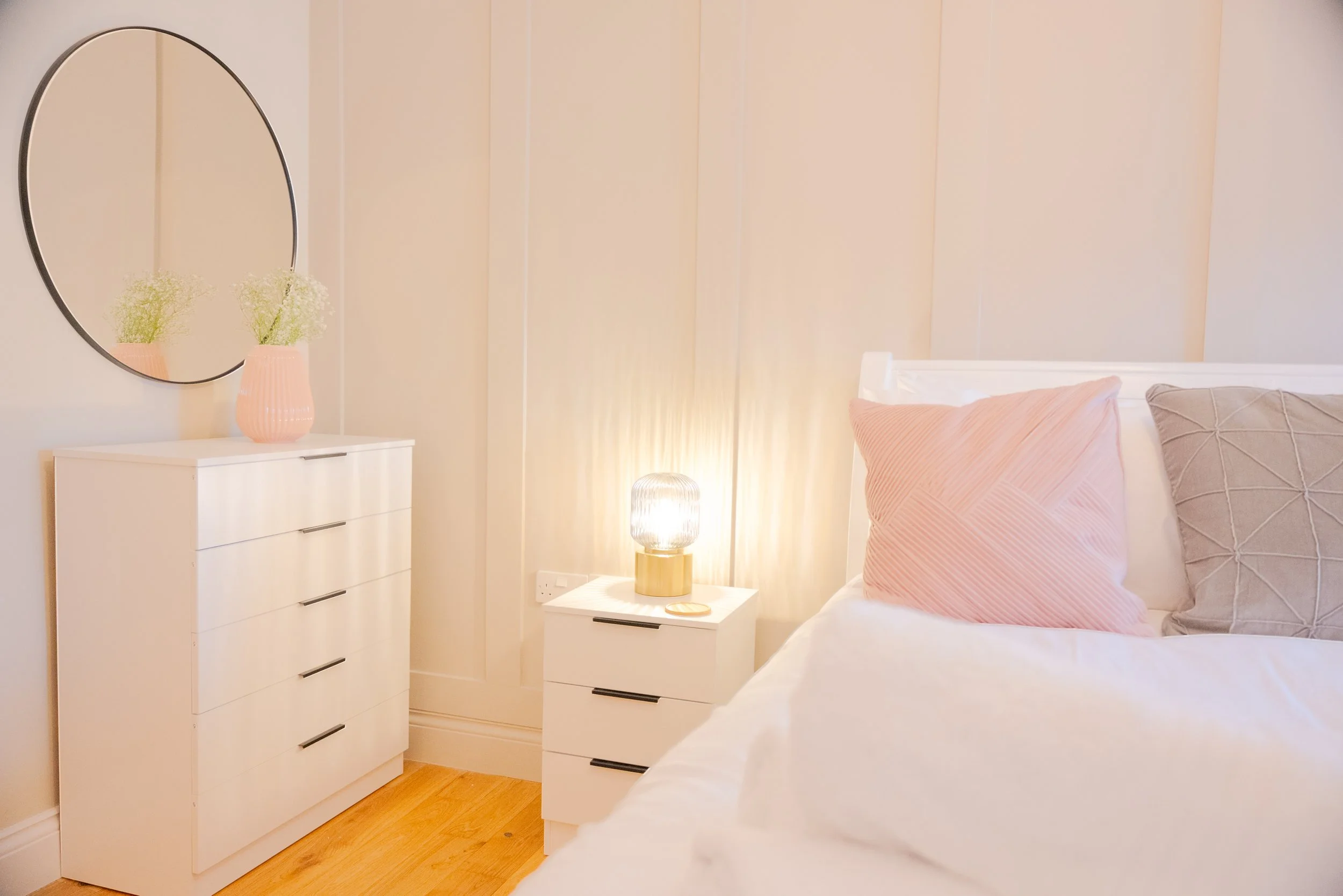 A cozy bedroom with a white dresser topped with a pink vase holding white flowers, a round mirror, a bedside table with a gold lamp, a bed with pastel pink and gray pillows, and soft lighting.
