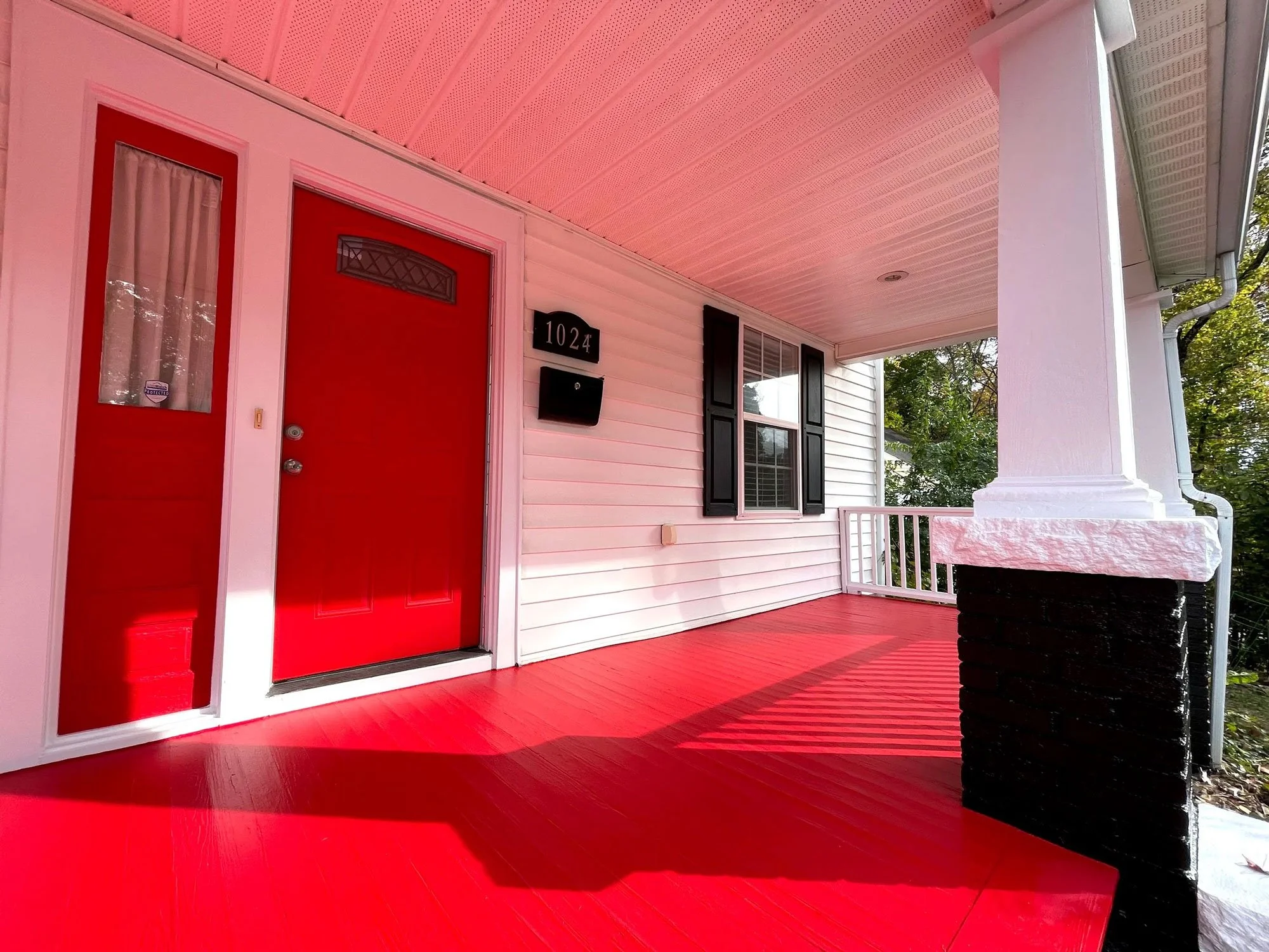 Front porch with red door, black mailbox, and black shutters on window, pink porch ceiling, red floor, white siding, surrounded by green trees.
