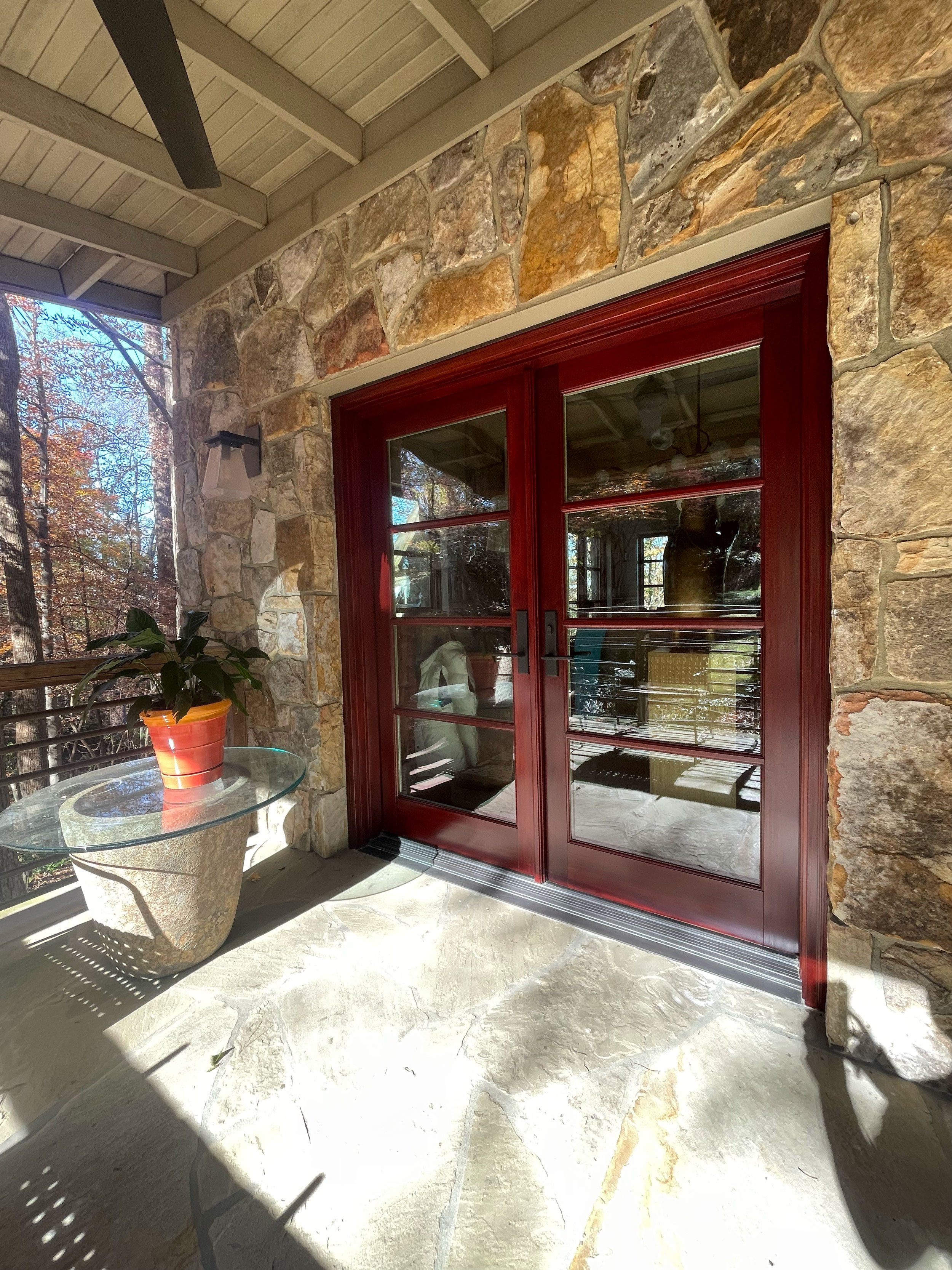 A porch area with a stone wall, red framed double glass doors, and a glass table with a potted plant. The porch is covered with a wooden ceiling and a wall-mounted light fixture. Trees with autumn leaves can be seen outside through the open railing.