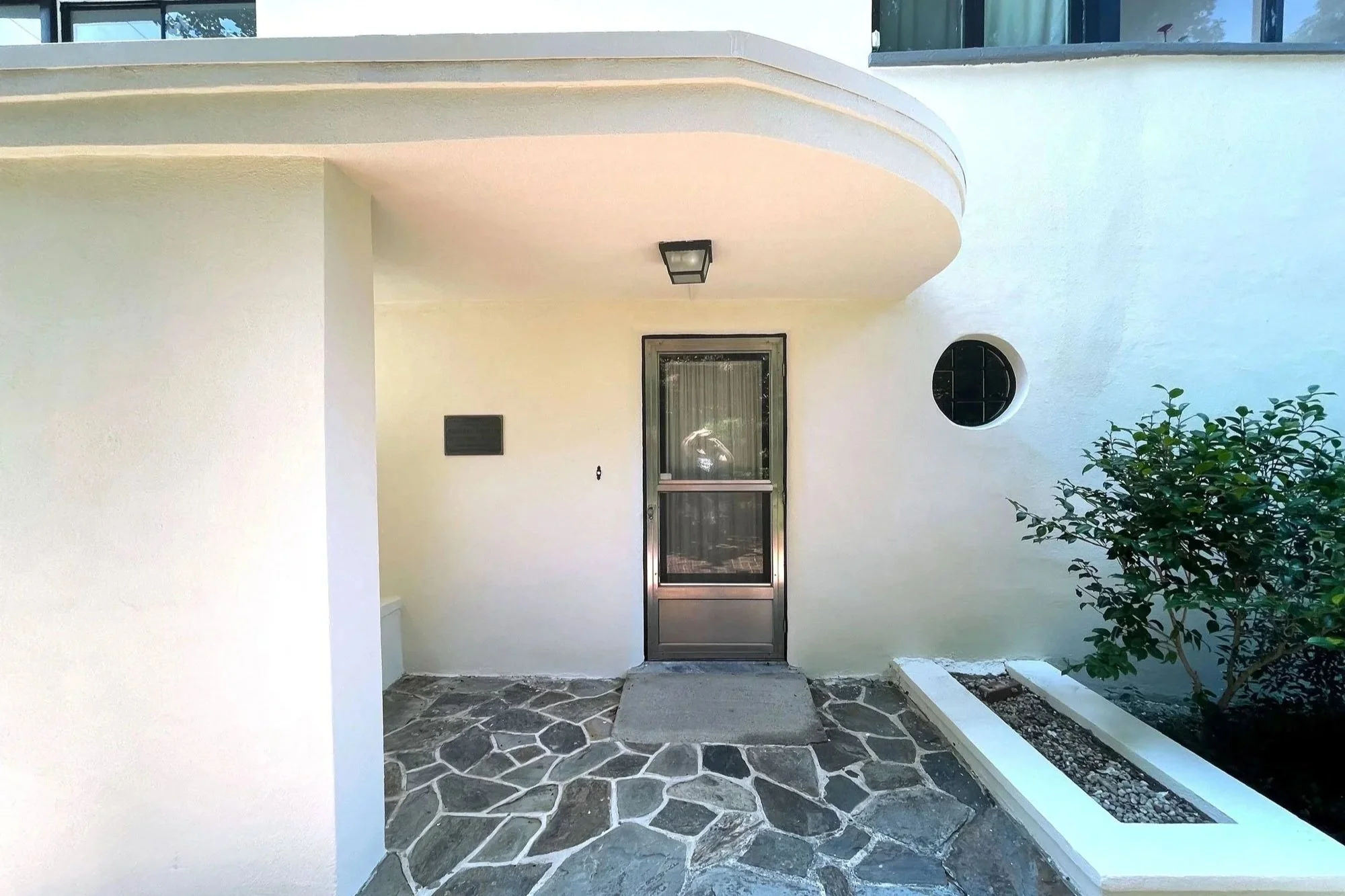 Front porch area of a house with a stone walkway, a glass door with a screen, a small window, and a side shrubbery.