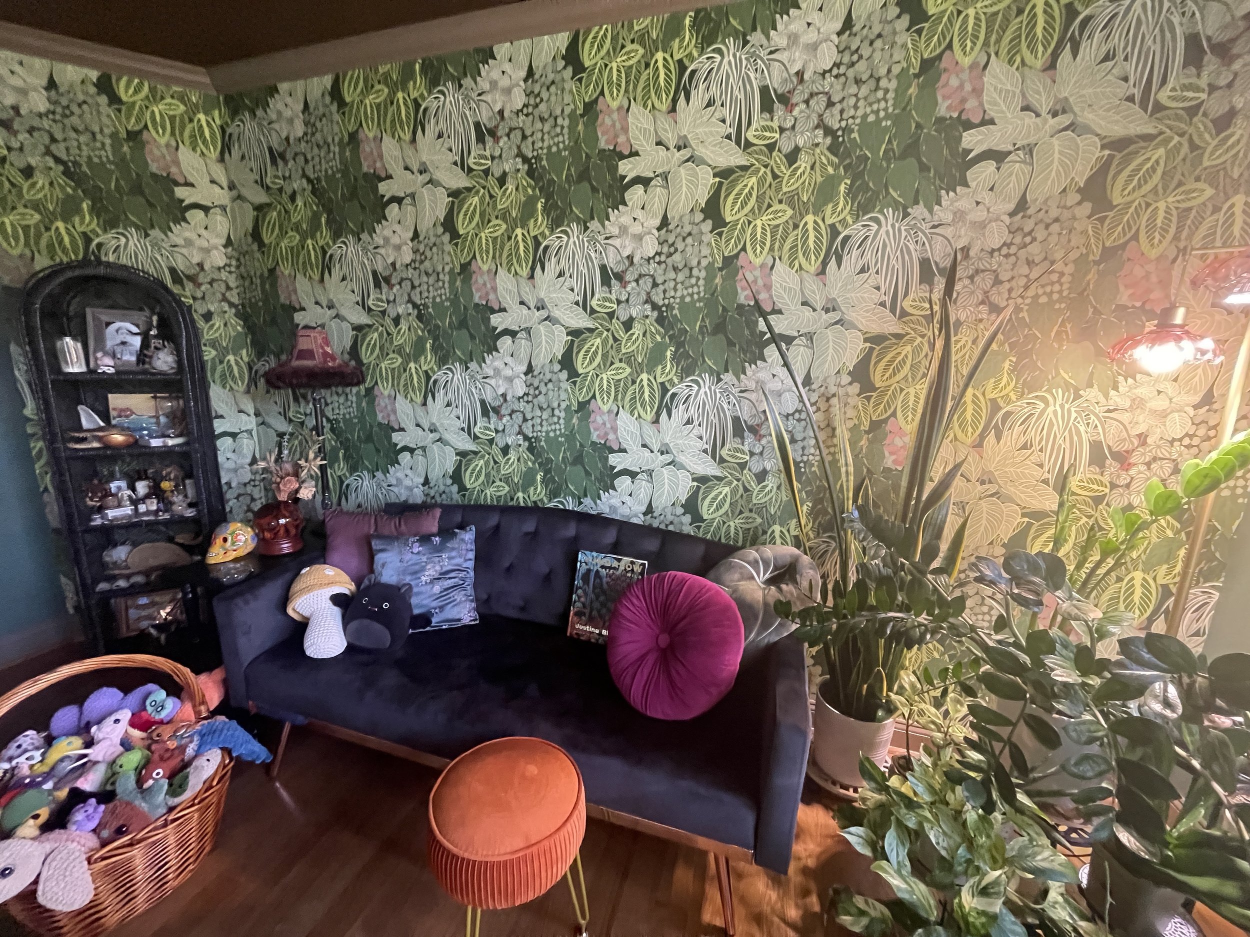 Living room with green leafy wallpaper, a black velvet couch decorated with pillows, an orange velvet stool, a black shelving unit with decorative items, a large indoor plant, and soft lighting from a lamp.