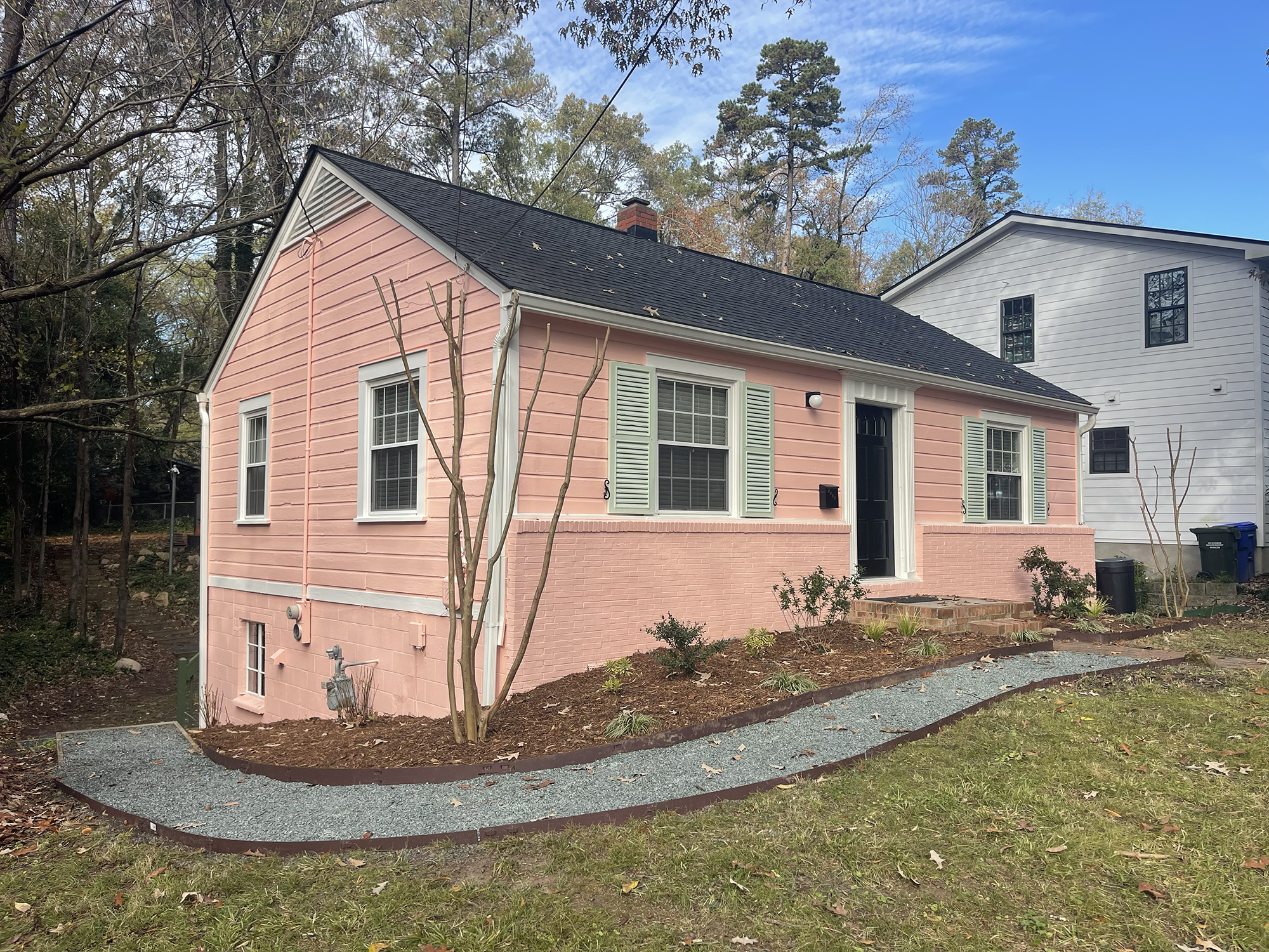 A pink house with white trim and a black roof, situated in a yard with small plants and mulch, surrounded by trees, under a partly cloudy sky.