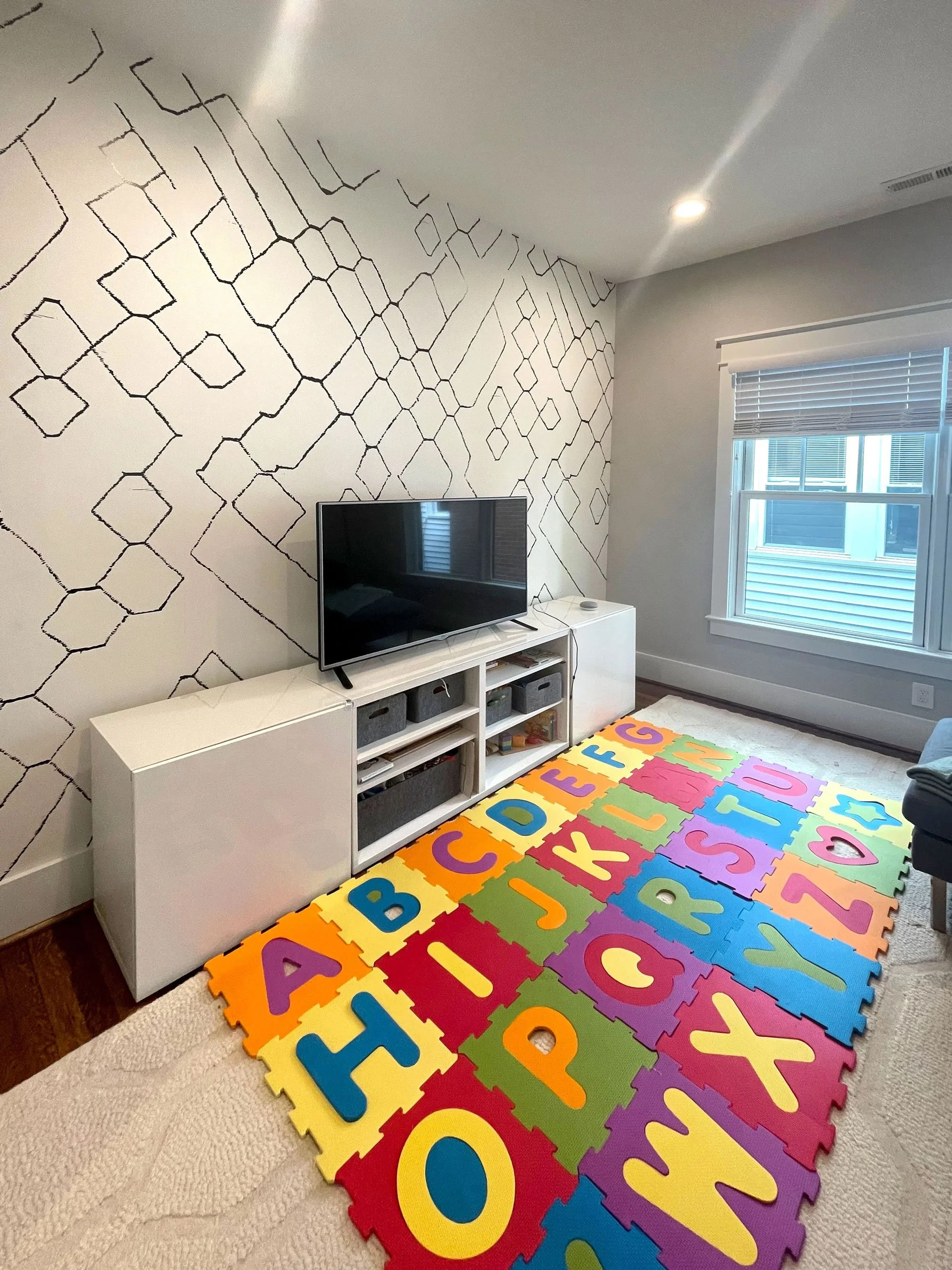 Colorful foam puzzle mat with alphabet letters in a child's playroom, with a TV on a white stand, a window with blinds, and a wall with black line art decor.