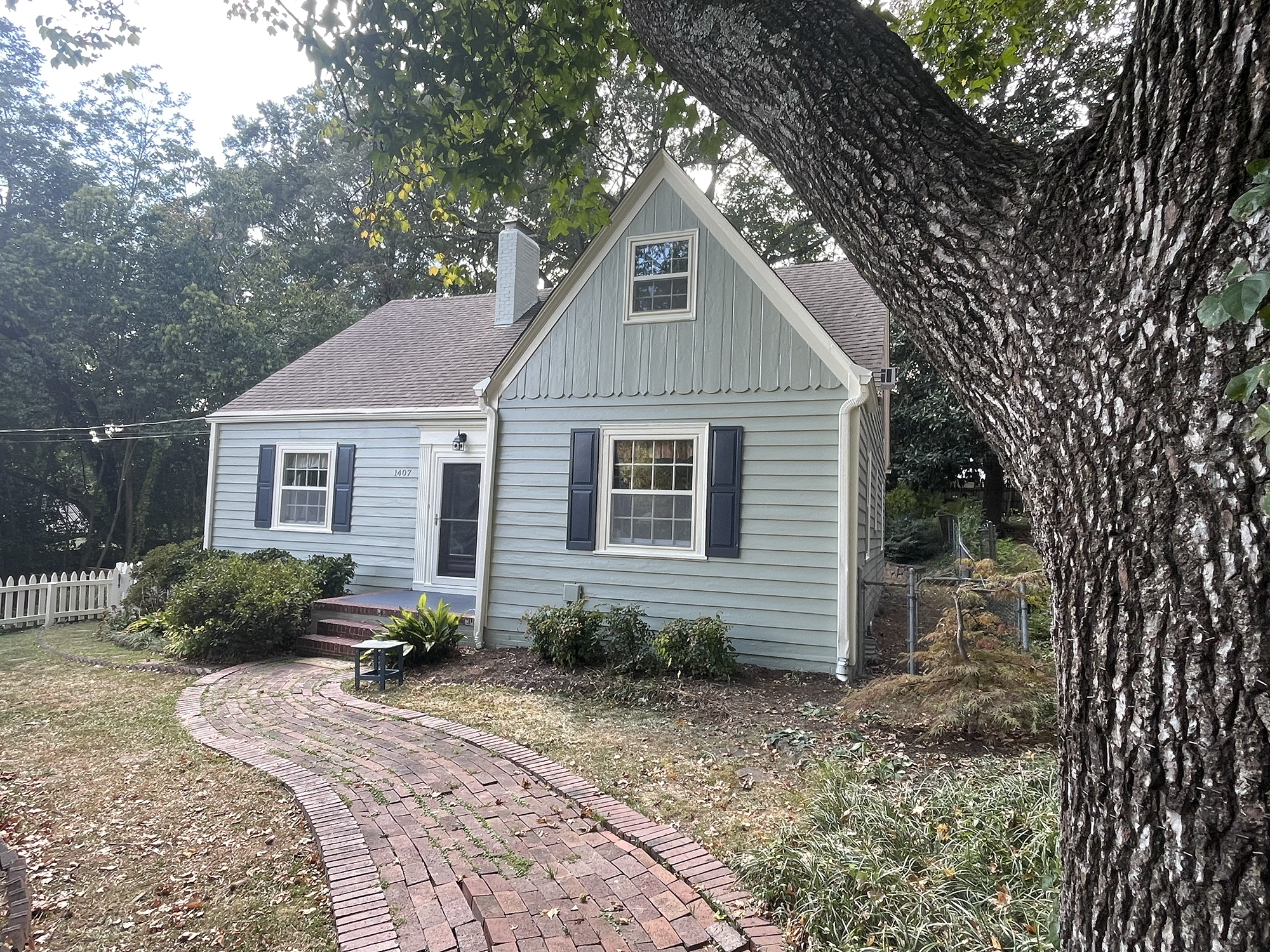 A house with light blue siding, dark blue shutters, and a brick pathway leading to the front door, surrounded by a yard and large tree with blossoming branches.