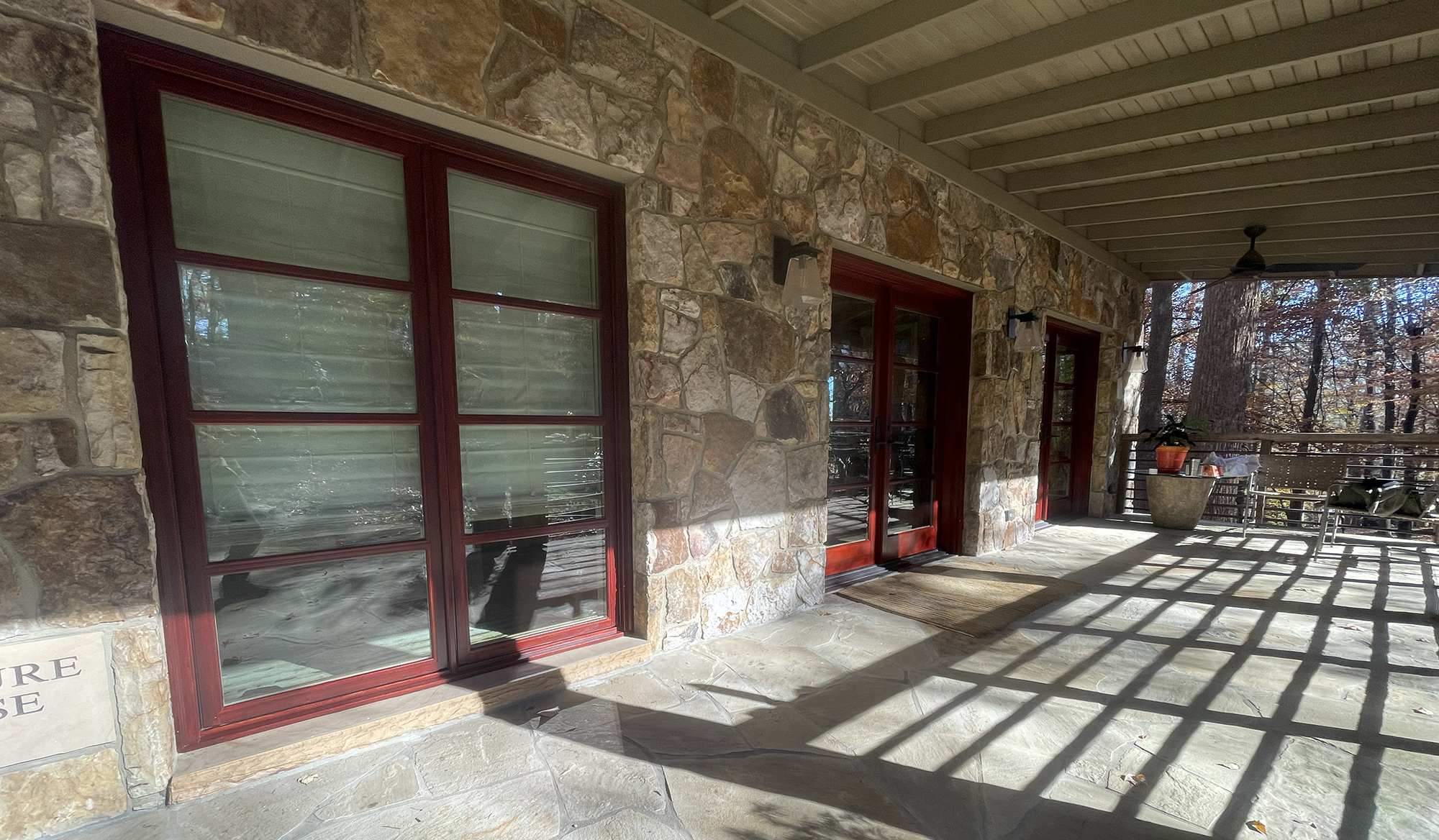 Stone porch with three red-framed glass doors, potted plants, and outdoor seating, surrounded by trees with fallen leaves.