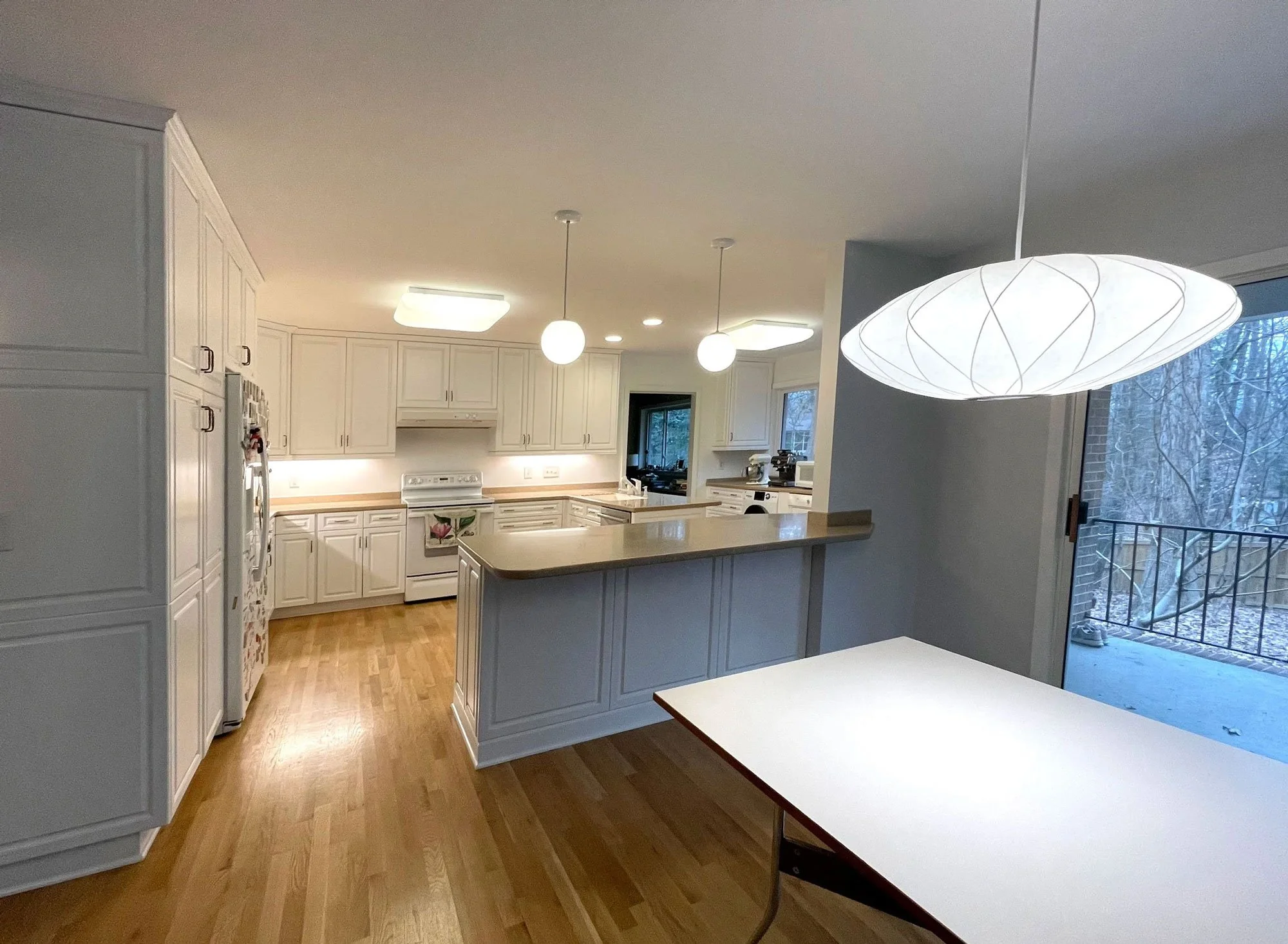 Kitchen with white cabinets, wooden flooring, island counter, modern lighting fixtures, and a large window showing trees outside.