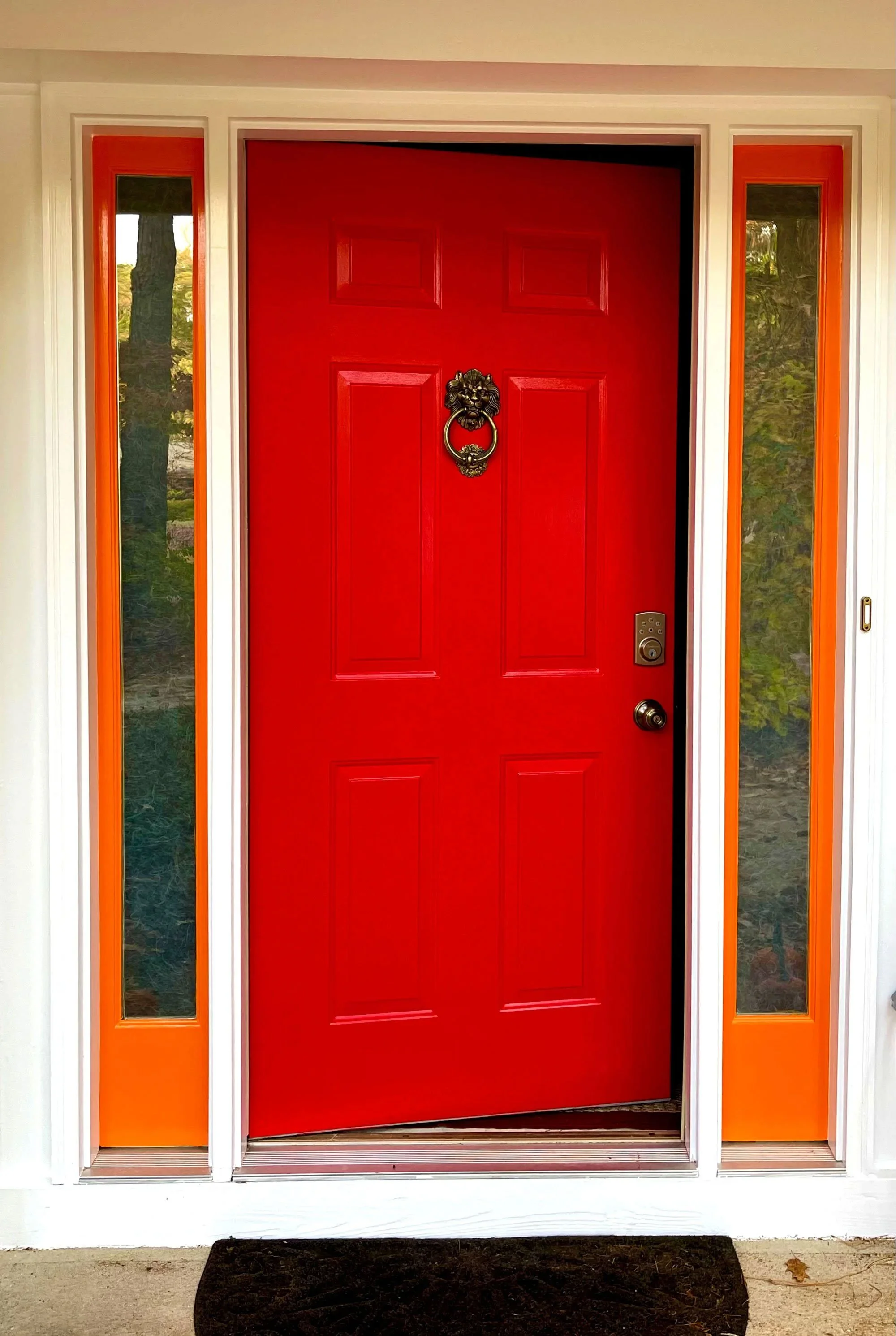 Red front door with lion head door knocker and electronic lock, flanked by two tall narrow windows with orange frames and glass panes, outside a house on a porch with a dark doormat and a fallen leaf.