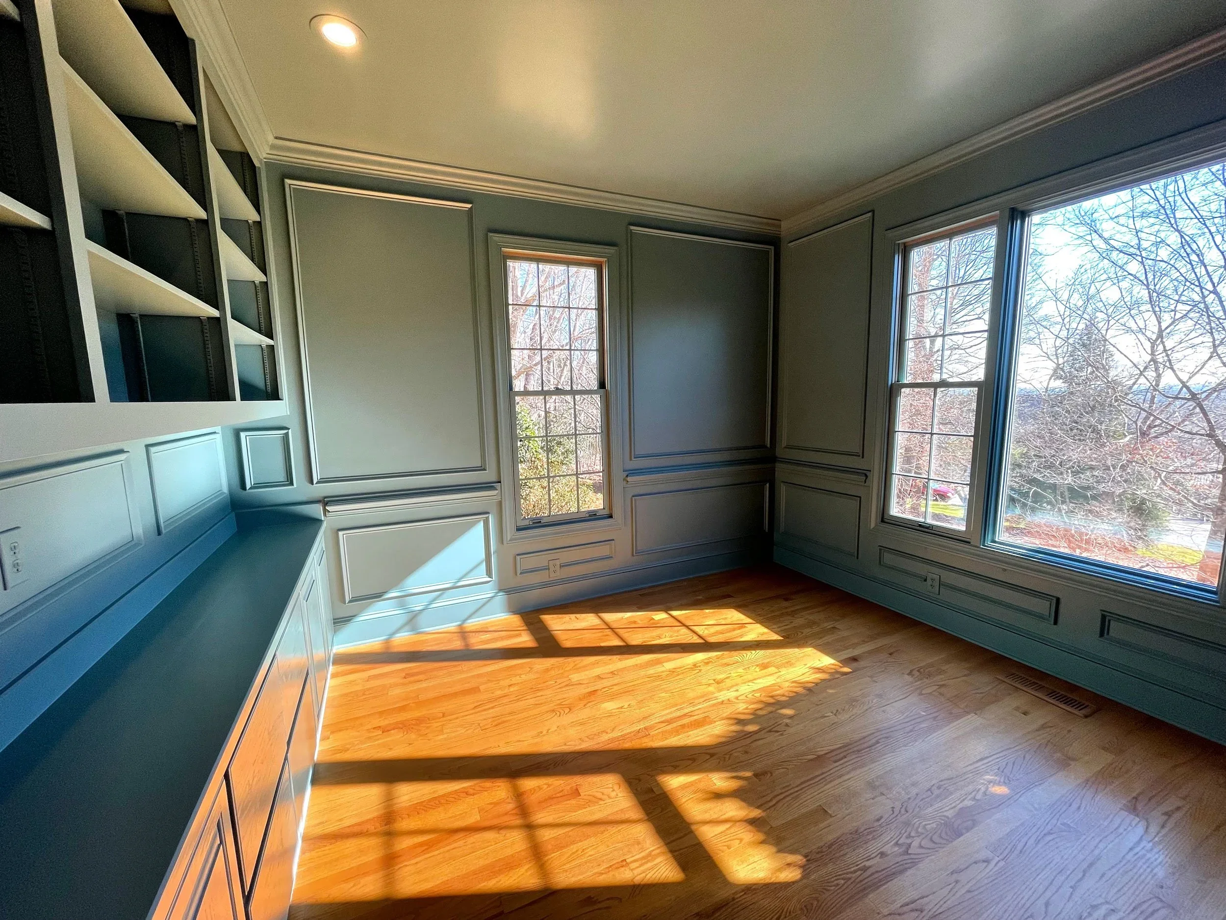 A room with wooden floor, large windows with trees outside, light-colored walls with decorative molding, a built-in bookshelf, and sunlight casting shadows on the floor.