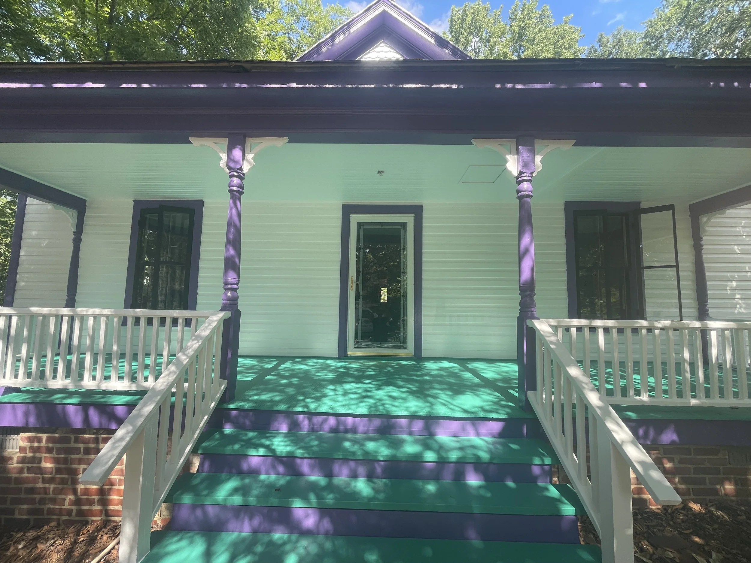 Front view of a house with a porch, white railing, purple porch columns, and a covered top. There is a green porch floor, a brick foundation, and trees and a blue sky in the background.