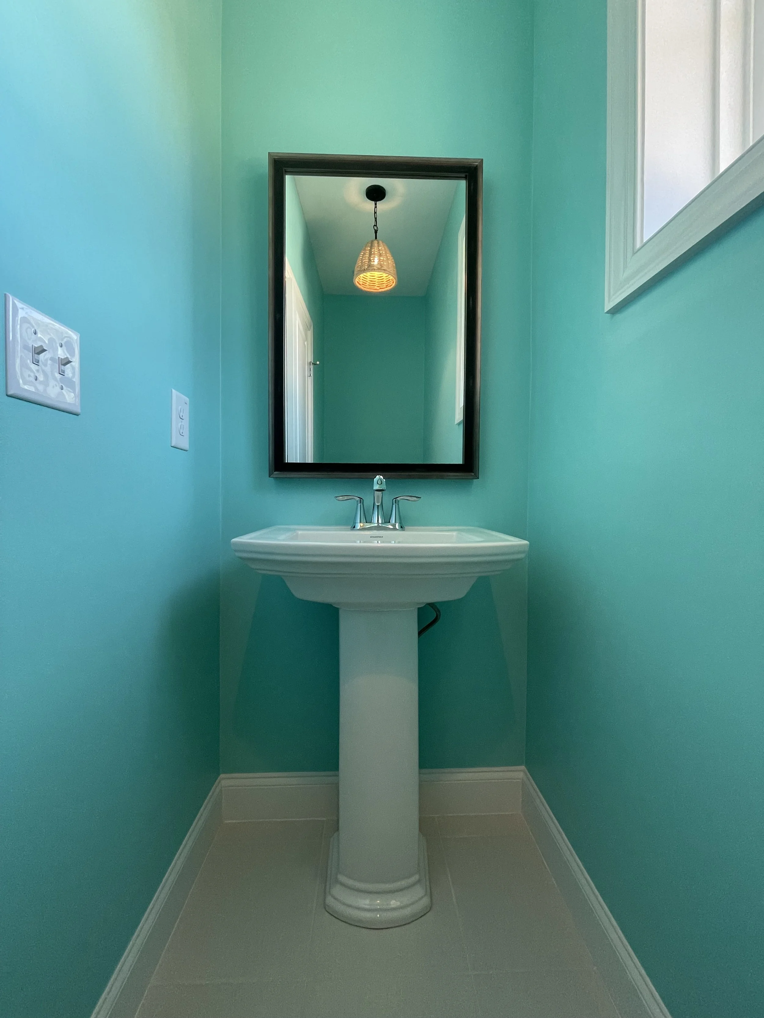 A spectacular bright teal powder room with a white pedestal sink, a black-framed mirror, a window with blinds, makes a small space sensational.
