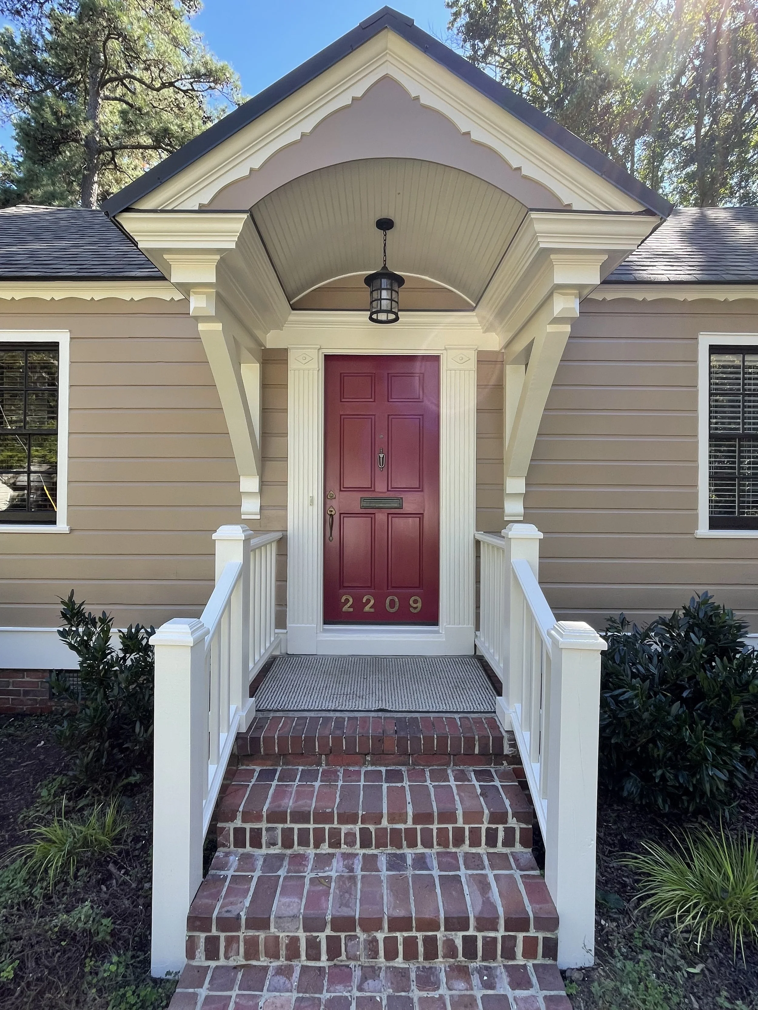 Front porch of a house with a red door, white columns, and brick stairs, surrounded by greenery and trees, under a sunny sky.