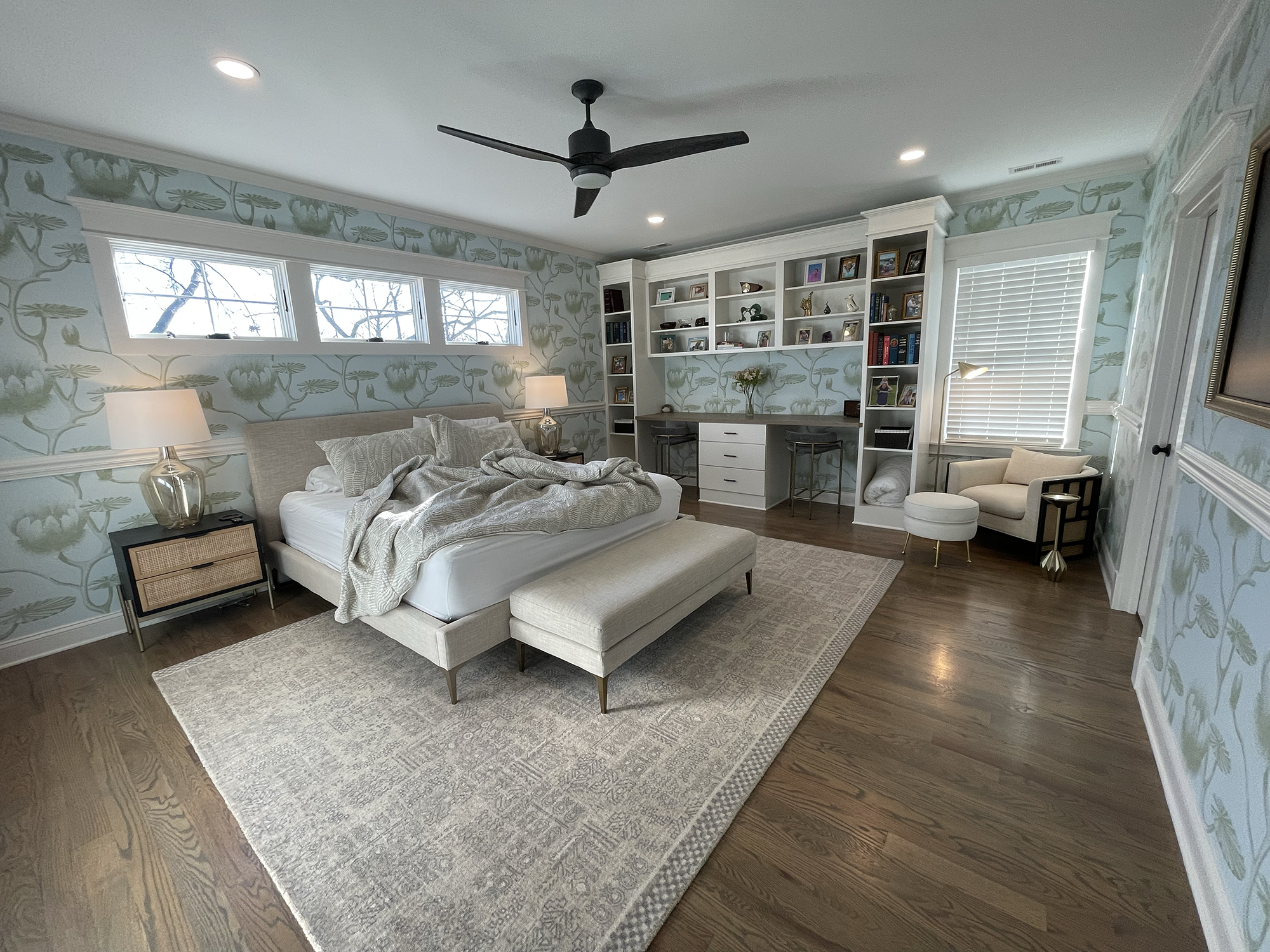 A spacious bedroom with light blue floral-patterned wallpaper, hardwood floors, a white bed with beige bedding, two nightstands with lamps, a large built-in white bookshelf with decorative items, small desk area with chairs, a window with blinds, and a ceiling fan.