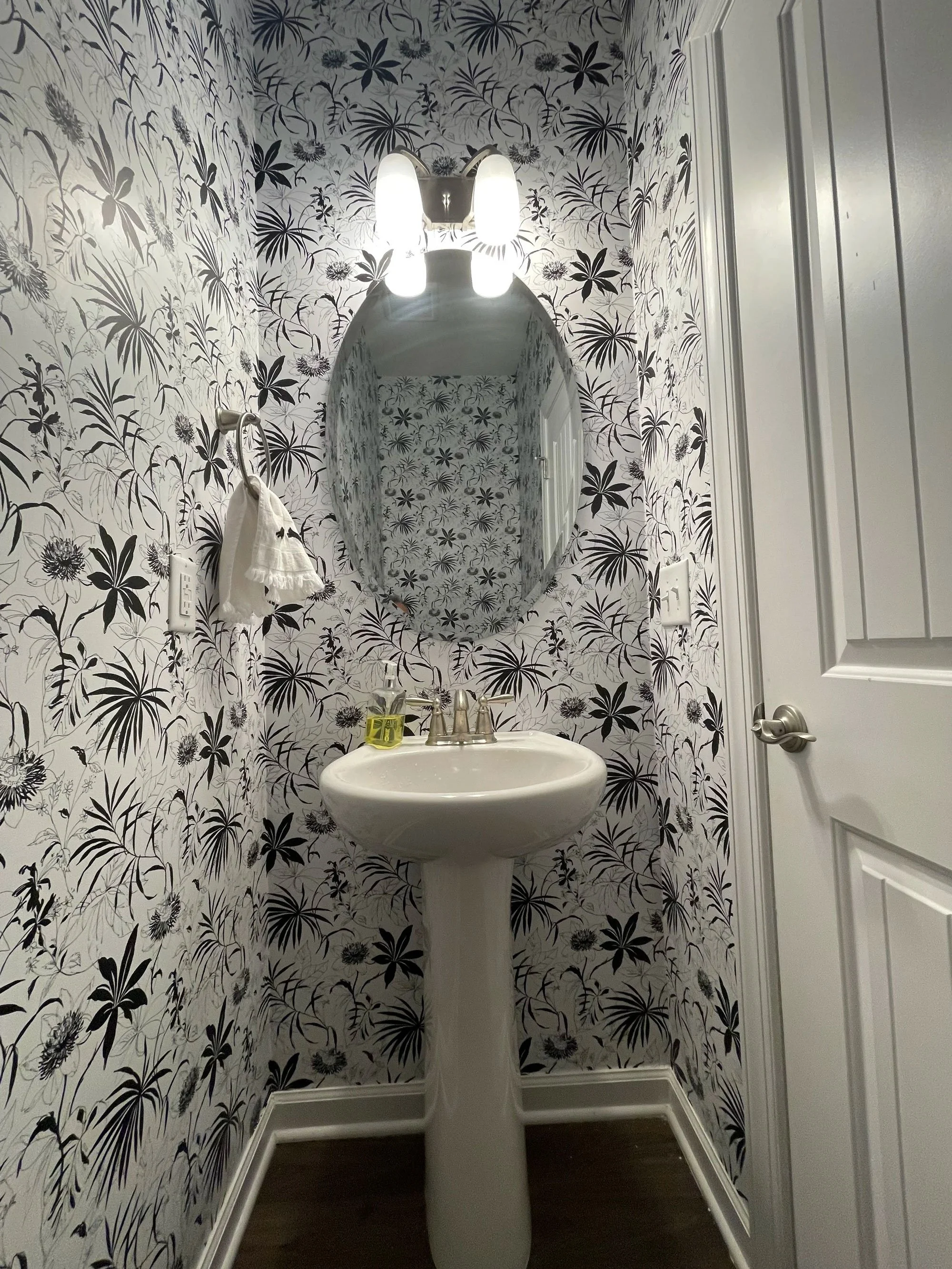 A bathroom with black and white floral wallpaper, a white pedestal sink with a silver faucet, a mirror, a soap dispenser, a towel on a towel ring, and closed white cabinet doors.