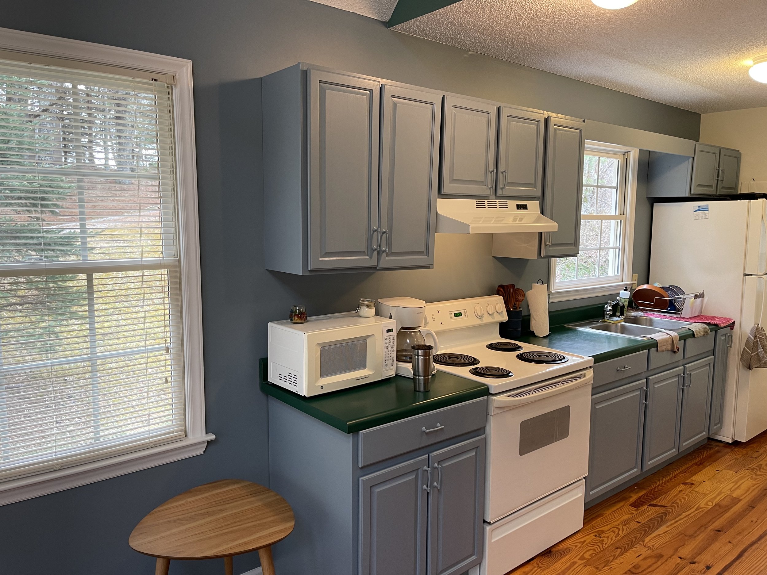 A kitchen with gray cabinets, a white stove, microwave, fridge, and two windows with blinds. There is a small wooden stool and various kitchen items on the counter.