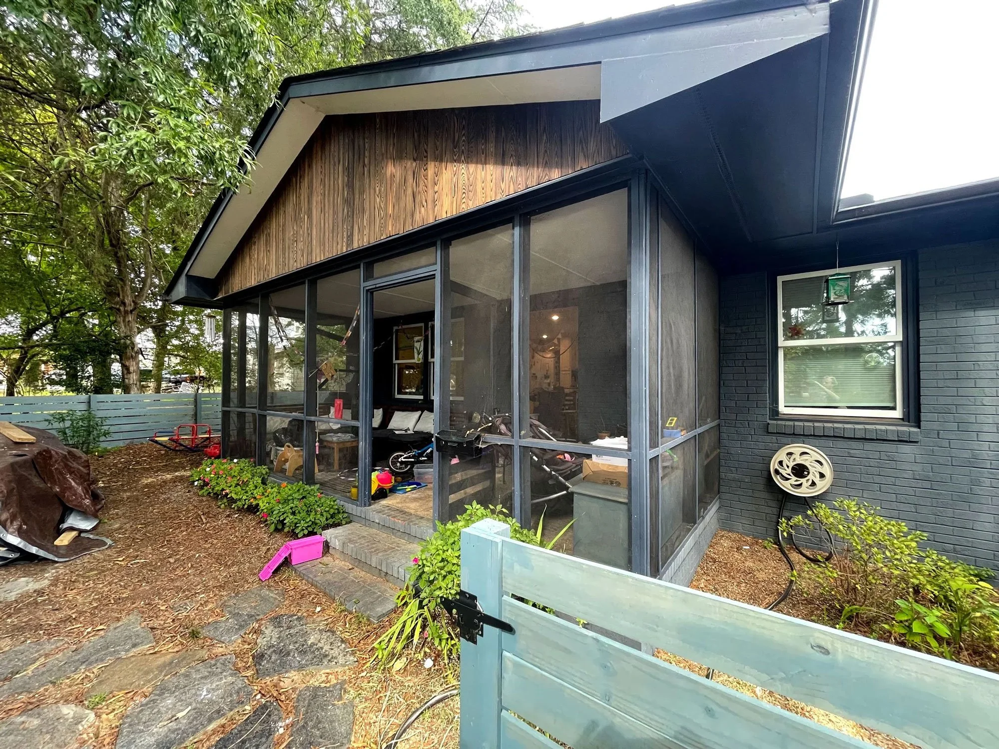 Backyard view of a house with a screened porch, green trees, a wooden fence, and various children's toys on the ground.