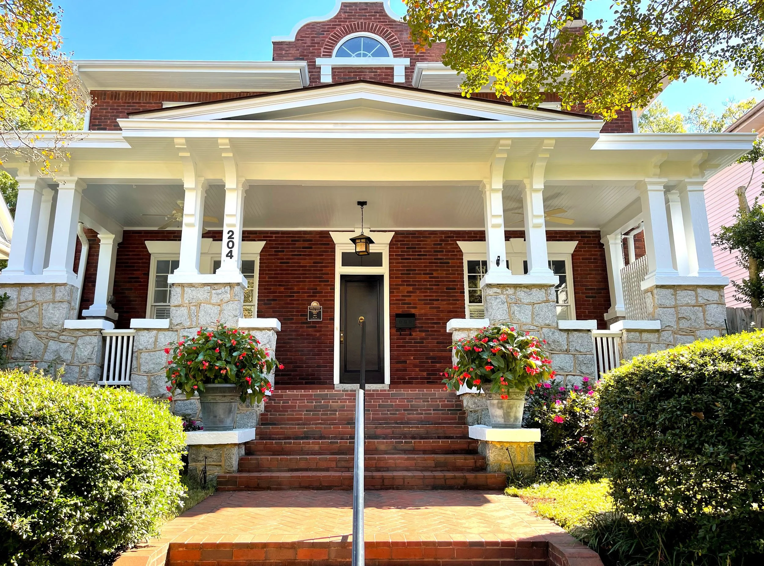A brick house with a front porch, white columns, and stairs leading up to the entrance. There are potted plants with flowers on either side of the stairs, and trees with green leaves framing the house. The house has a gabled roof and a small window above the porch.