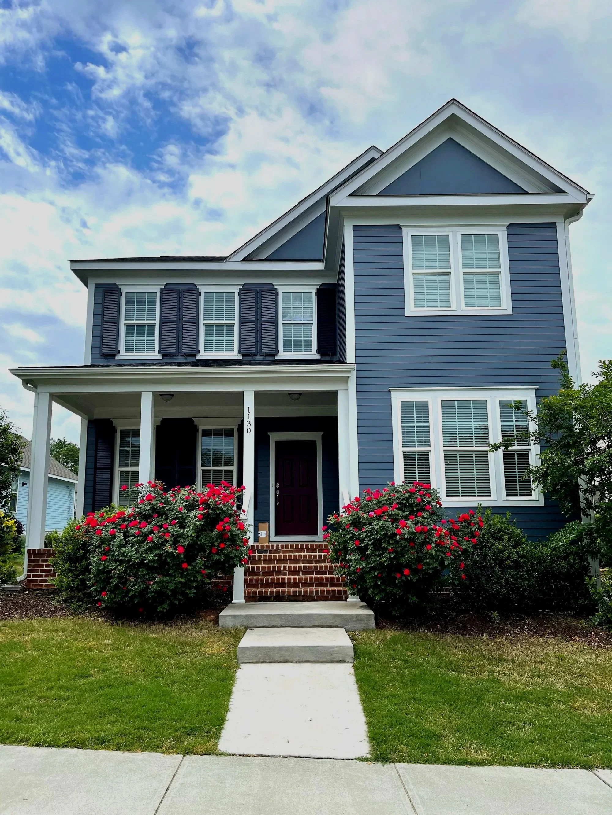 A two-story blue house with white trim, a covered front porch, and a stairway leading to the front door. The house has multiple windows with white shutters. There are red and pink flowers on either side of the bottom of the stairs, with green grass and a sidewalk in front. The sky is partly cloudy with blue sky visible.