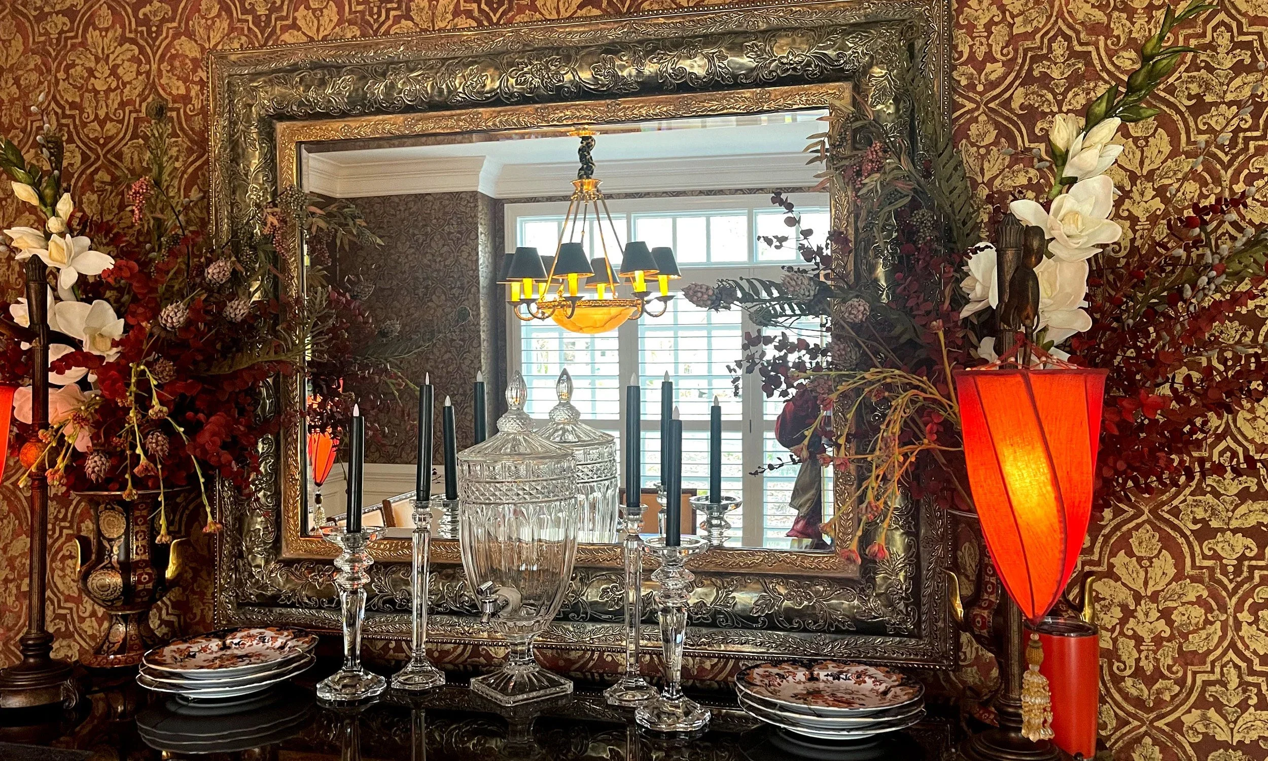 A decorative interior with a large ornate silver mirror, a chandelier, floral arrangements, black candlesticks, glassware, plates, and a red lampshade. The room features patterned wallpaper and a window with blinds.