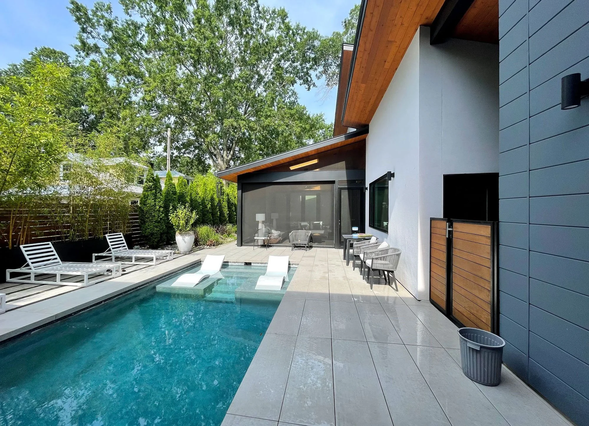 Modern backyard with a swimming pool, lounge chairs, and outdoor seating, surrounded by trees and greenery.