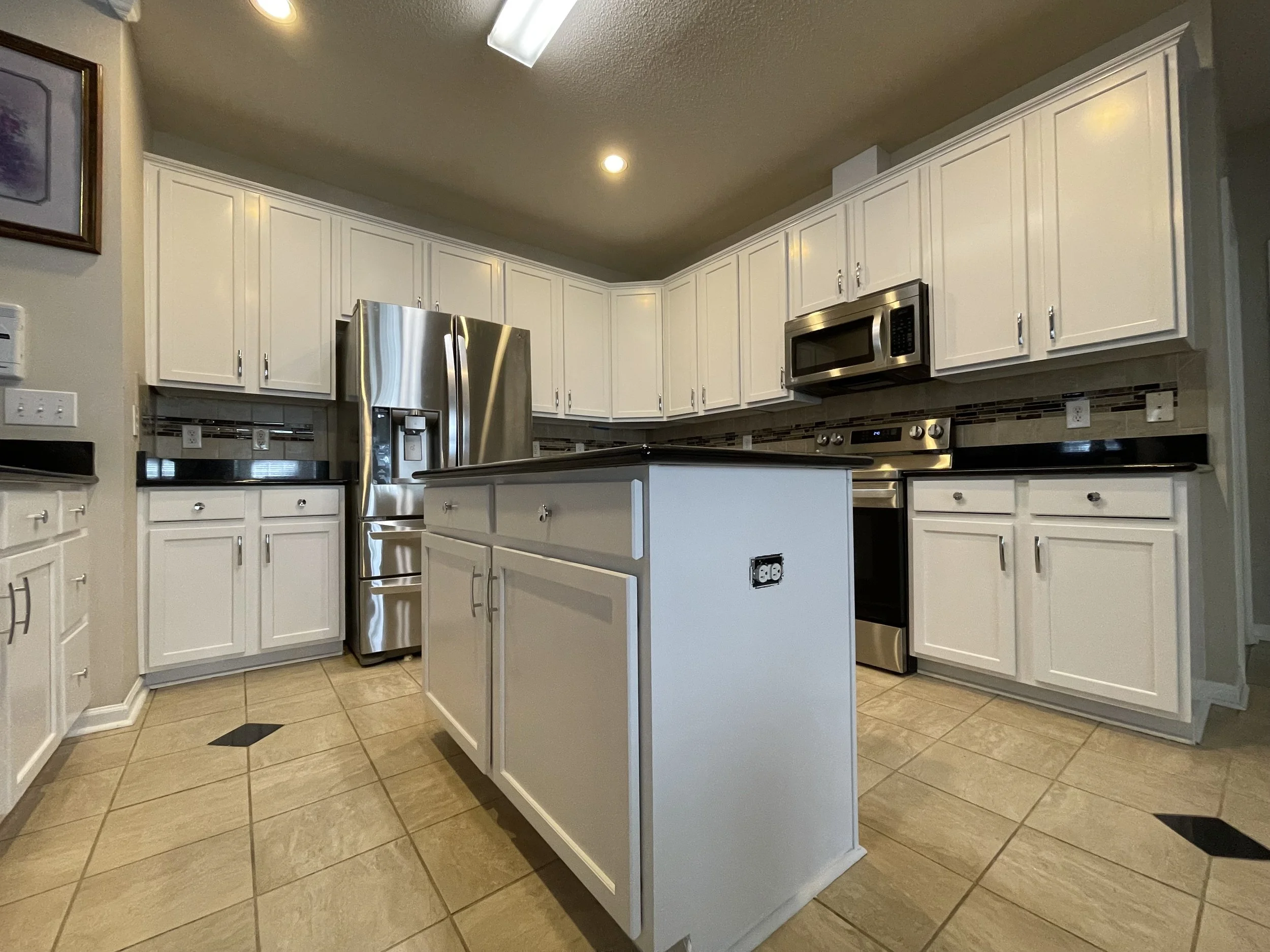 Kitchen with white cabinets, stainless steel refrigerator, microwave, and oven. Small kitchen island, tile floor, and partial wall with artwork.