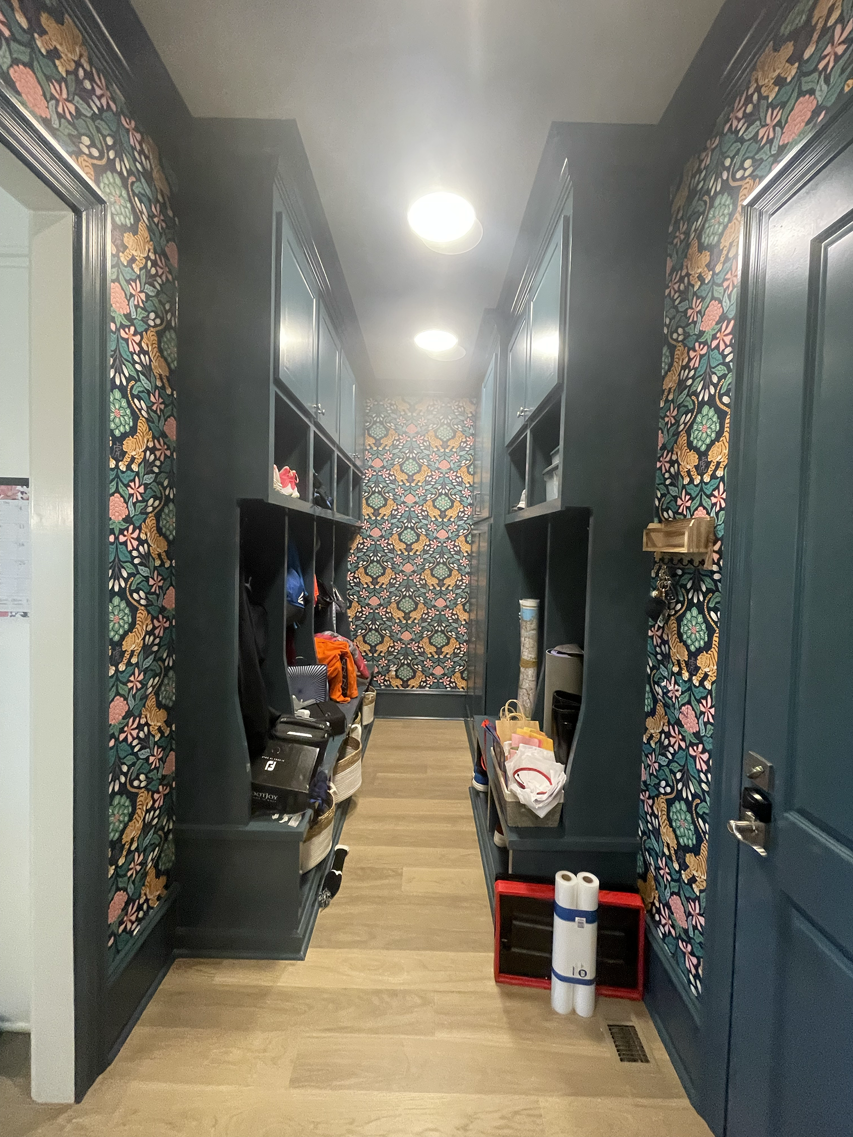 A narrow hallway with dark blue walls and floral wallpaper border. Black cubbies with personal items and shoes line both sides. The floor is wood, and the ceiling has three recessed lights. Cleaning supplies and paper towels are on the floor at the end of the hallway.