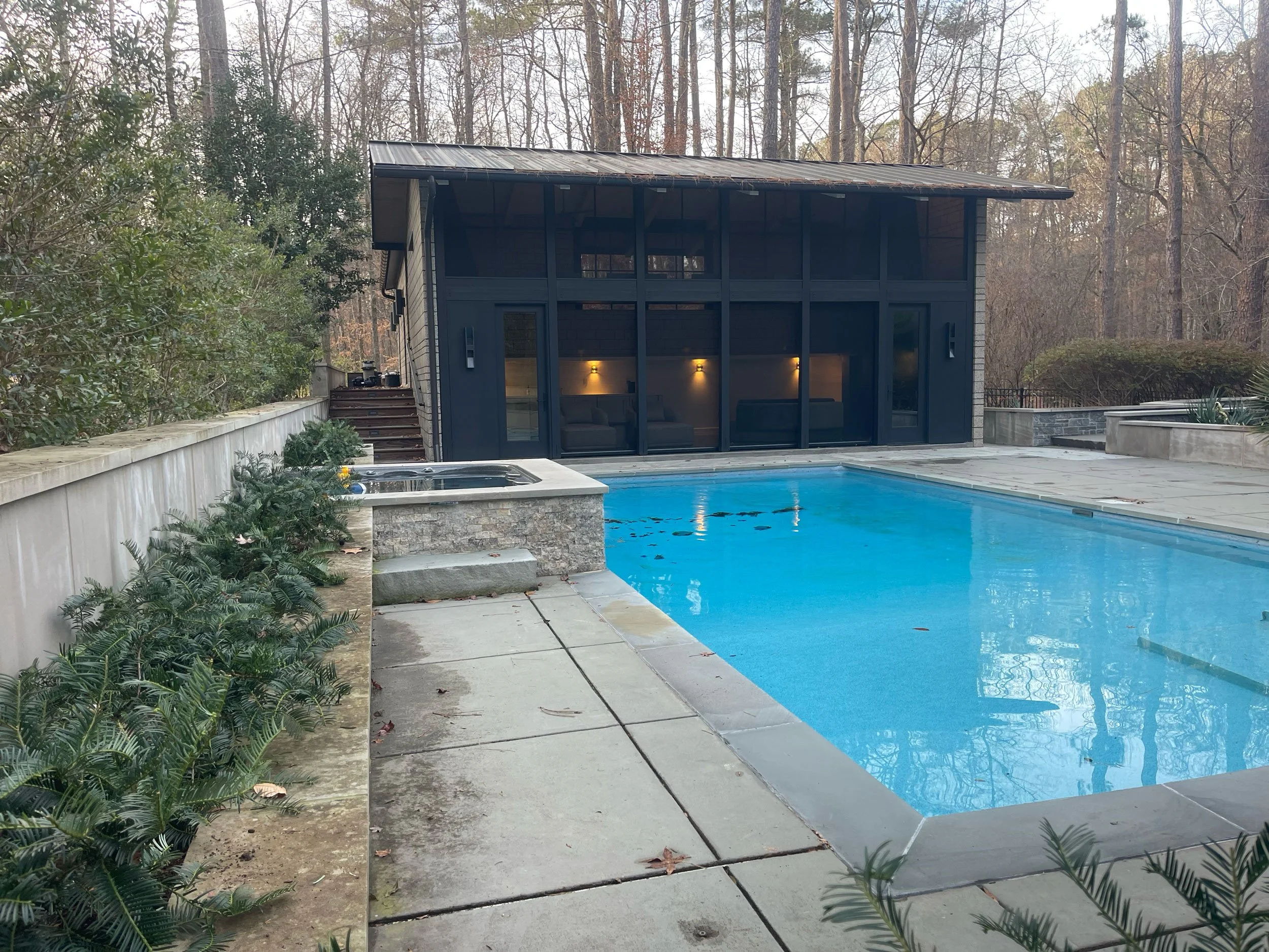 A custom designed pool house with a swimming pool, surrounded by a concrete deck and lush greenery, houses a kitchen, a fireplace, a shower and a sauna for the best of times.