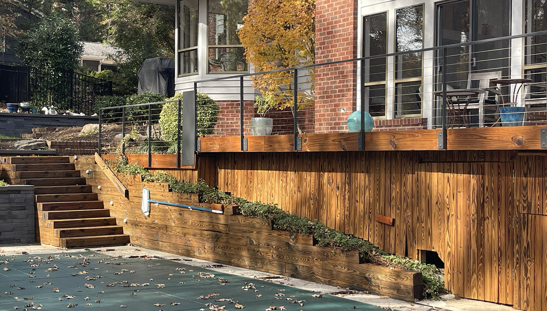 A multi-level backyard with stairs, a pool with fallen leaves, and a wooden deck with outdoor furniture and decorative items, adjacent to a brick house with large windows.