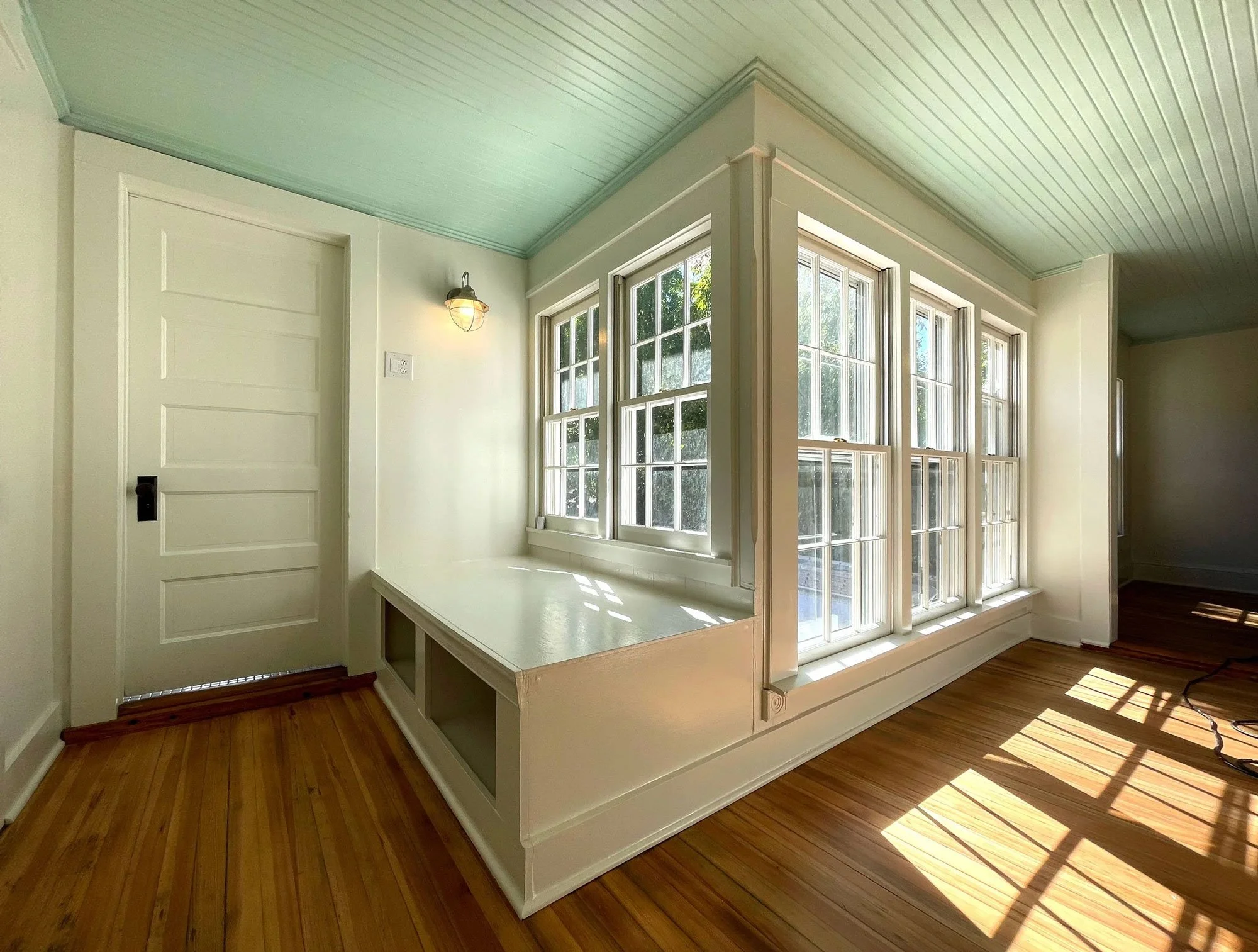 A sunroom with large windows, wooden floor, white walls, and a built-in bench. Sunlight casts shadows on the floor.