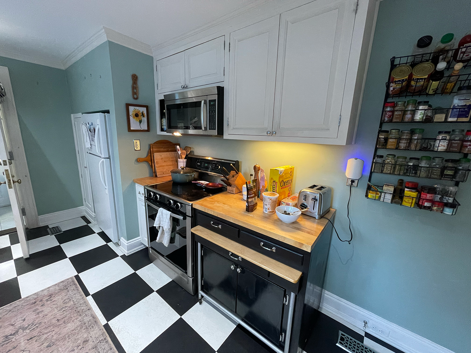 A kitchen with checkered black and white flooring, a stove with a pot on it, wooden countertop with cereal box, bowls, toaster, and various food items, white cabinets, a mounted microwave, spice rack on the wall, and a small lamp plugged into an outlet.