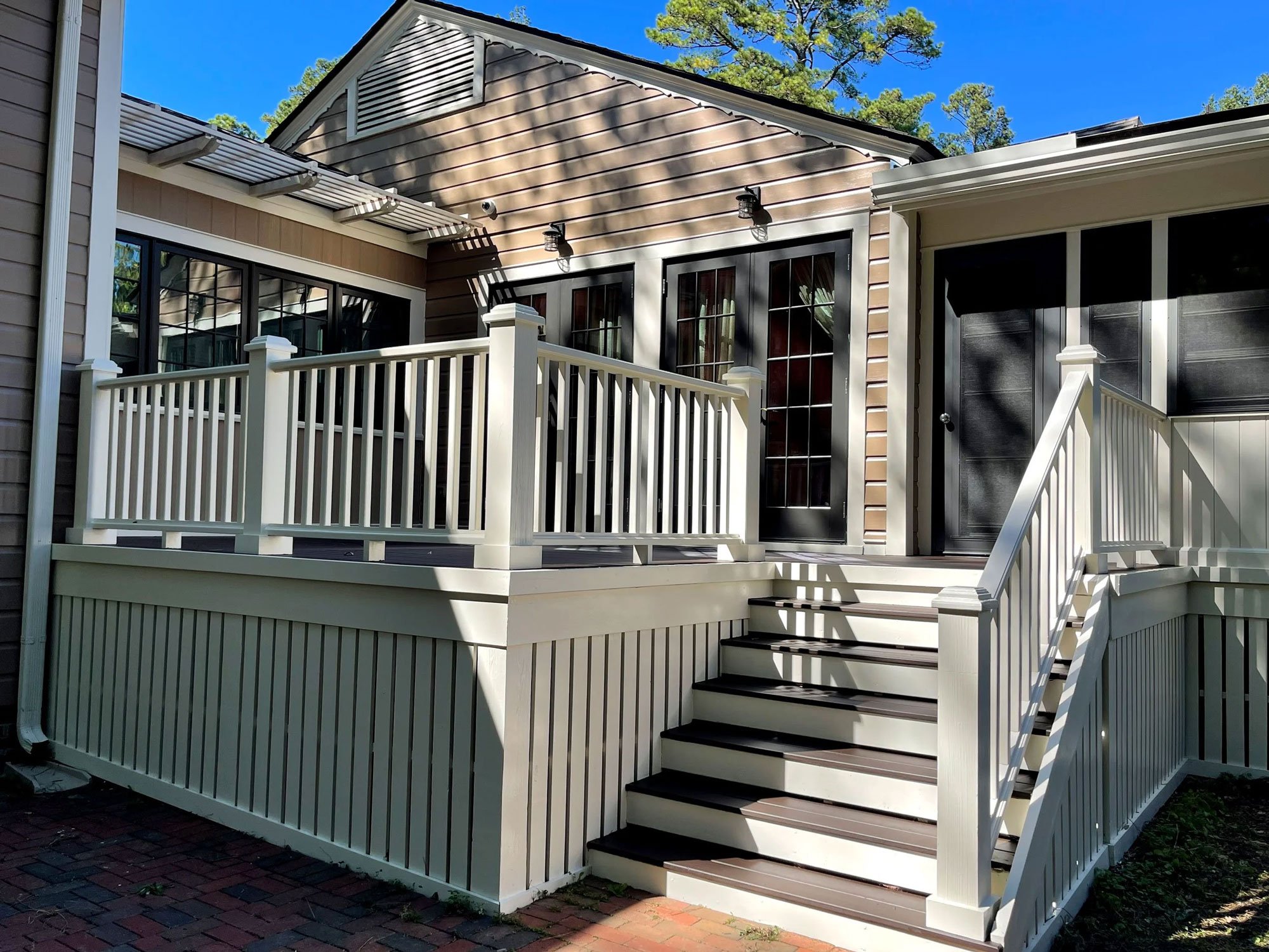 A white wooden staircase with black treads leading to a porch of a house. The house has light brown siding, a black-framed glass door, and black-framed window panes. There are two outdoor wall lamps above the door. The scene is sunny with shadows of nearby trees cast on the house, and the sky is clear blue.