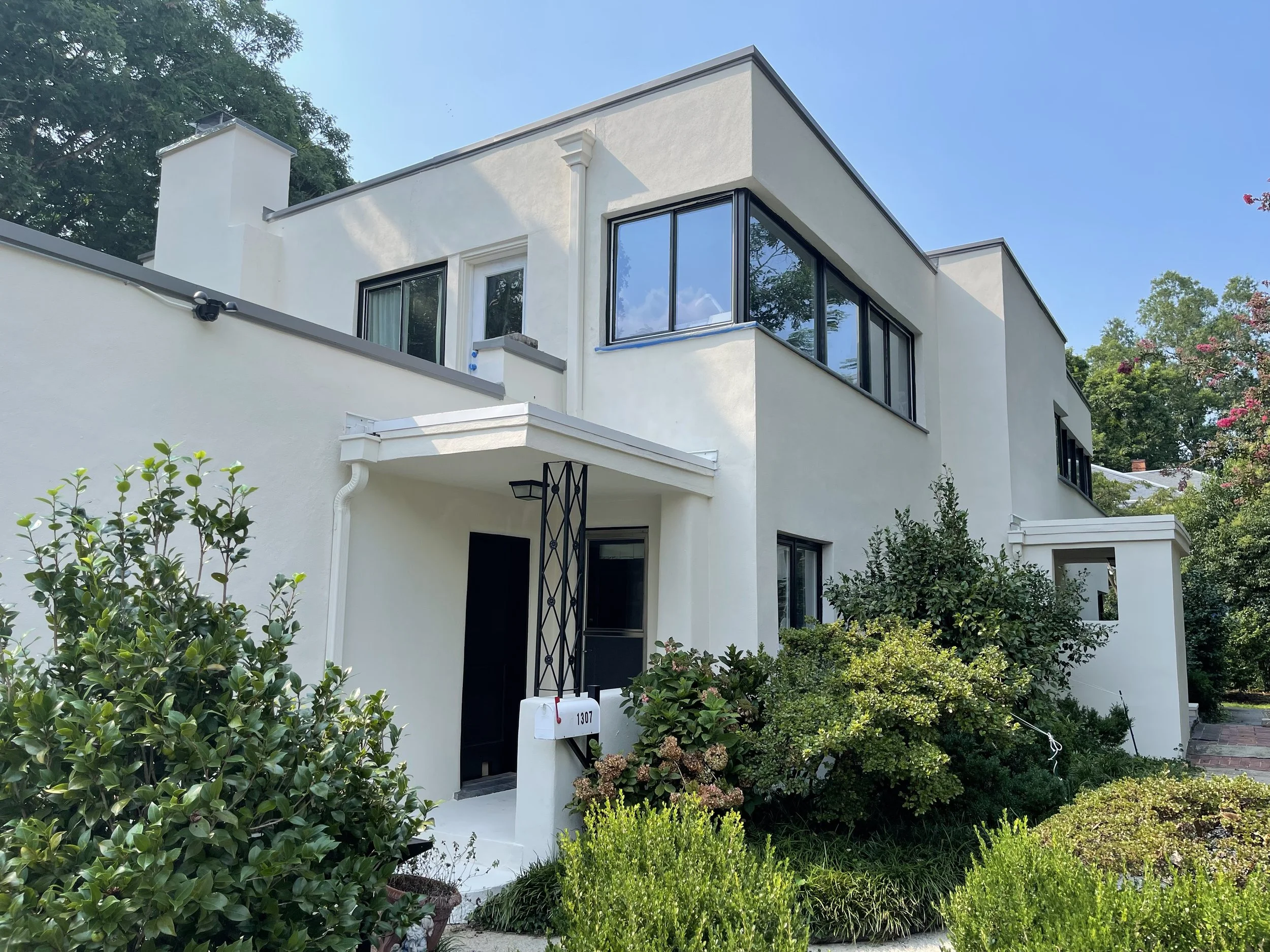 White modern house with large black-framed windows, surrounded by green bushes and plants, under a clear blue sky.