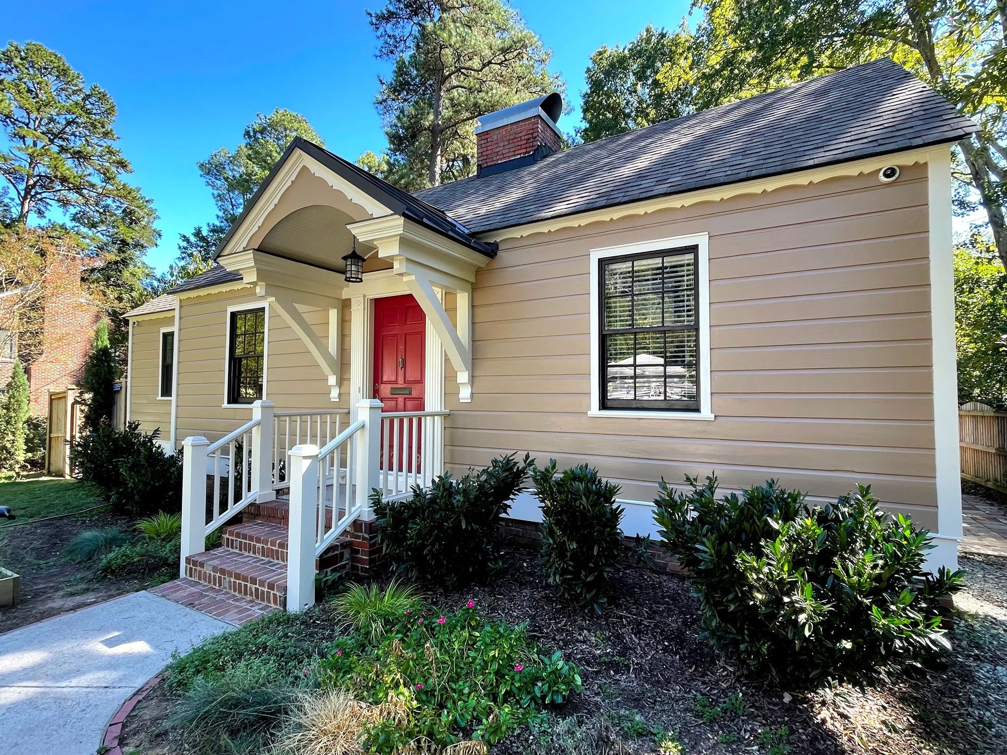 Front view of a beige house with red door, white porch railing, and brick steps, surrounded by a landscaped garden with bushes, small plants, and a concrete walkway, with tall trees and a clear blue sky in the background.