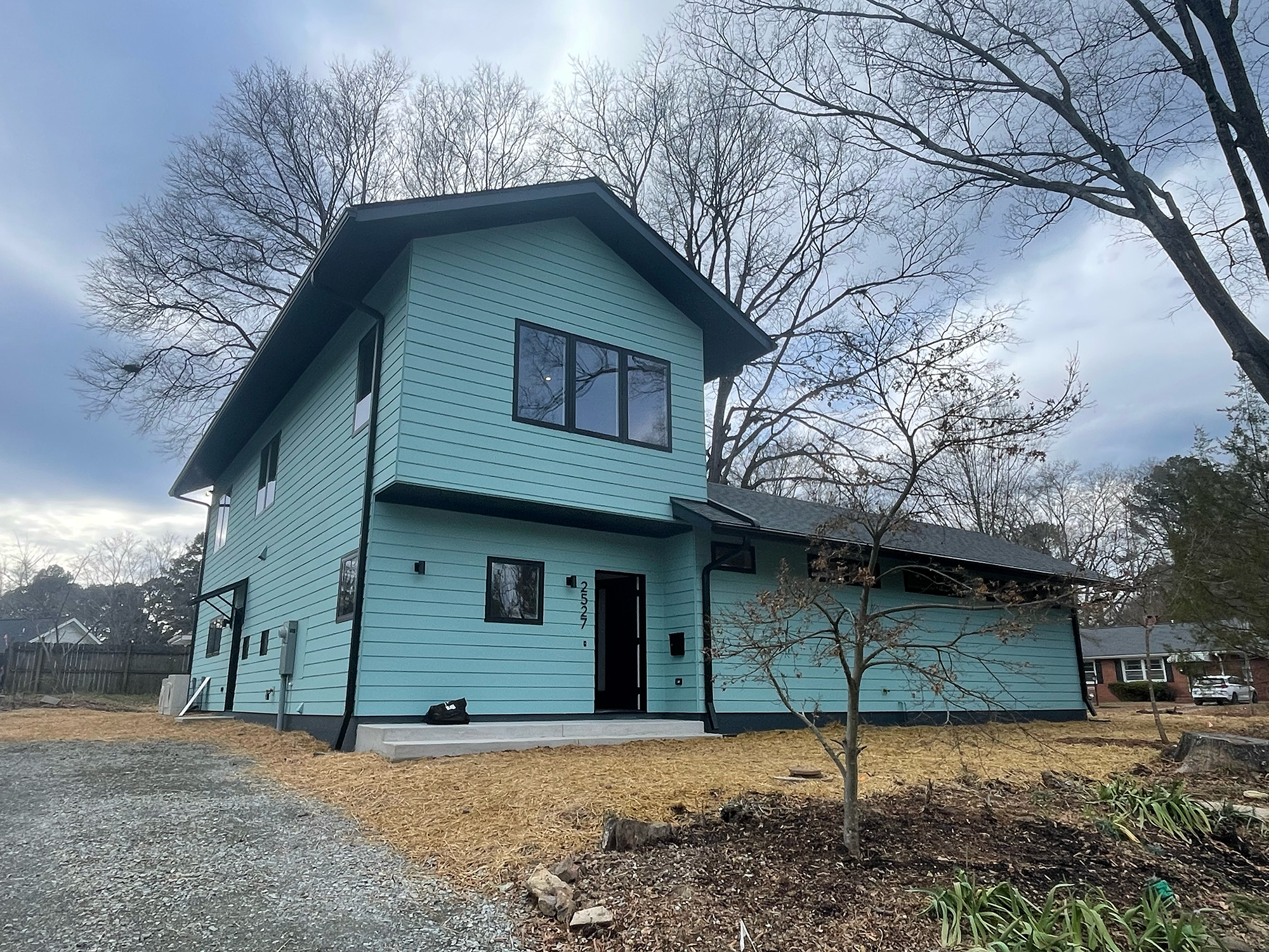 A light blue multi-story house with black window frames, a gabled roof, and a small porch. There are leafless trees in the background and a gravel pathway in the front yard.