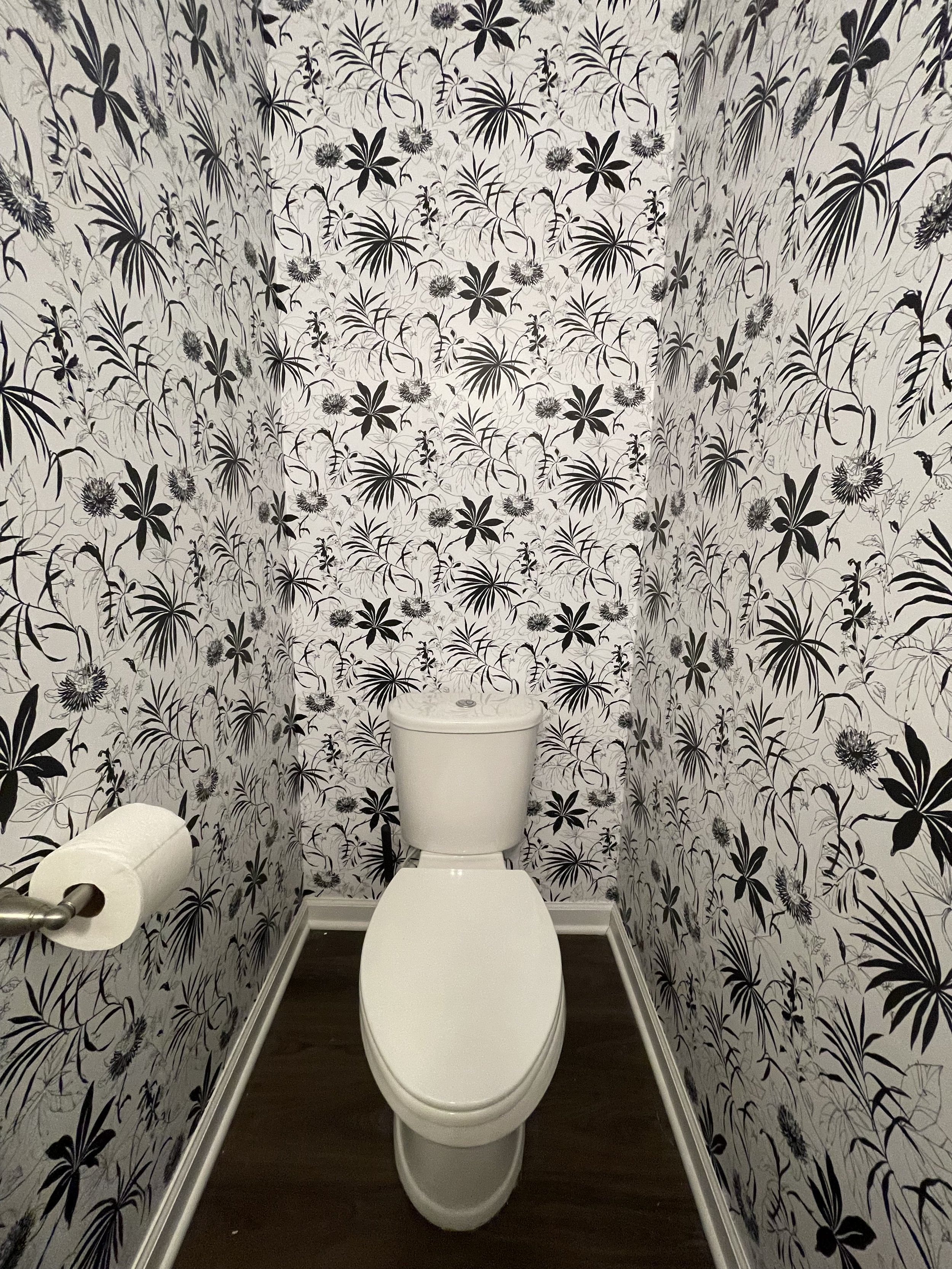A small bathroom with floral black and white patterned wallpaper, a white toilet, and a toilet paper roll on a holder on the left side.