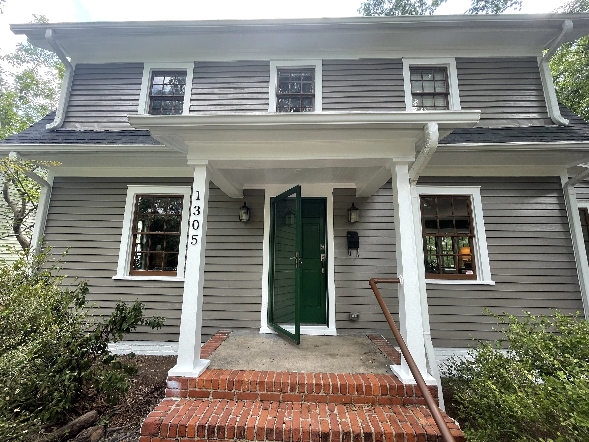 Front view of a two-story house with gray siding, white trim, and a green front door. There are three windows on the upper level and two on the lower level. The house has a small porch with brick stairs and a white support beam. Green trees and foliage are visible around the house.
