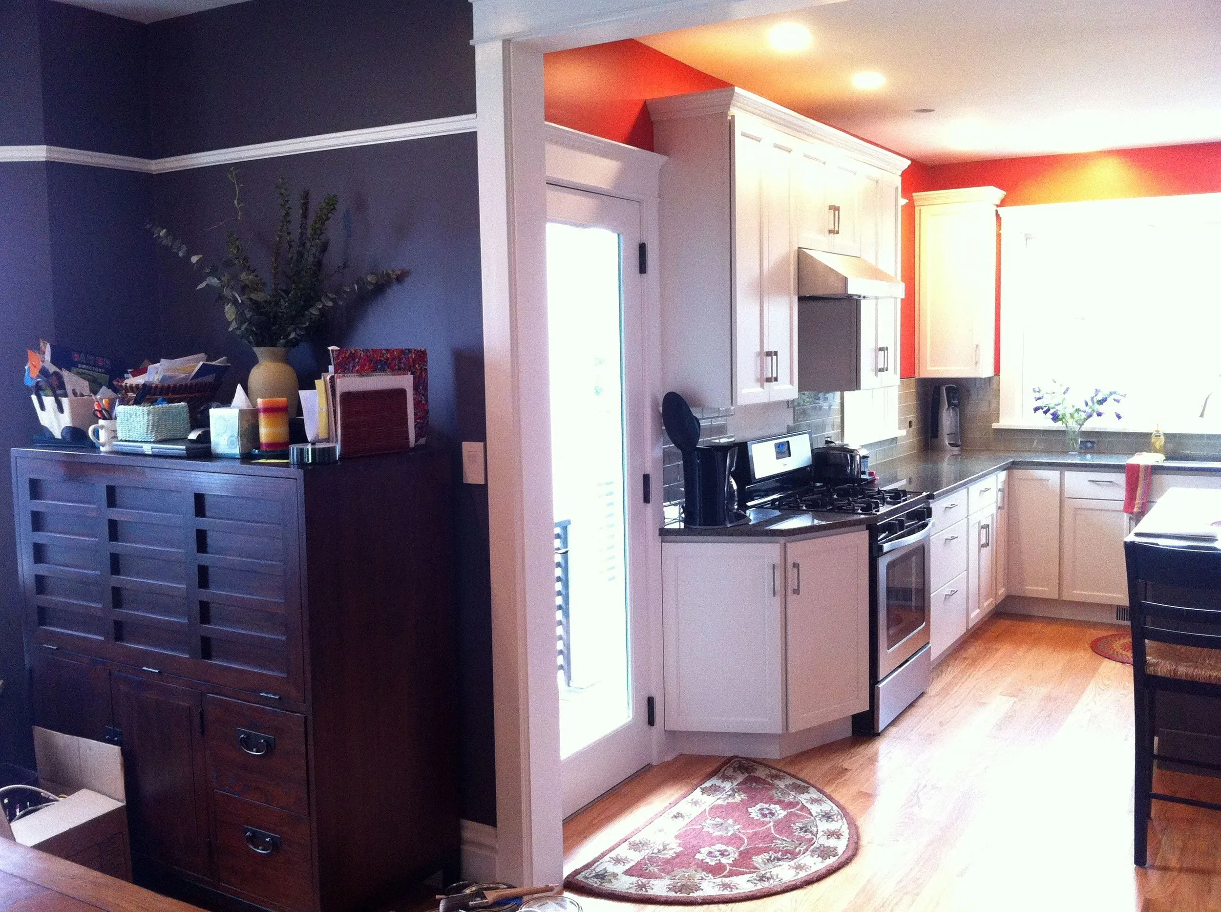 A kitchen with white cabinets, a stainless steel stove, and a black countertop. There is a small window with bright light, a potted plant on the countertop, and a red and beige patterned rug on the wooden floor. Light-colored walls with red accent walls are visible.