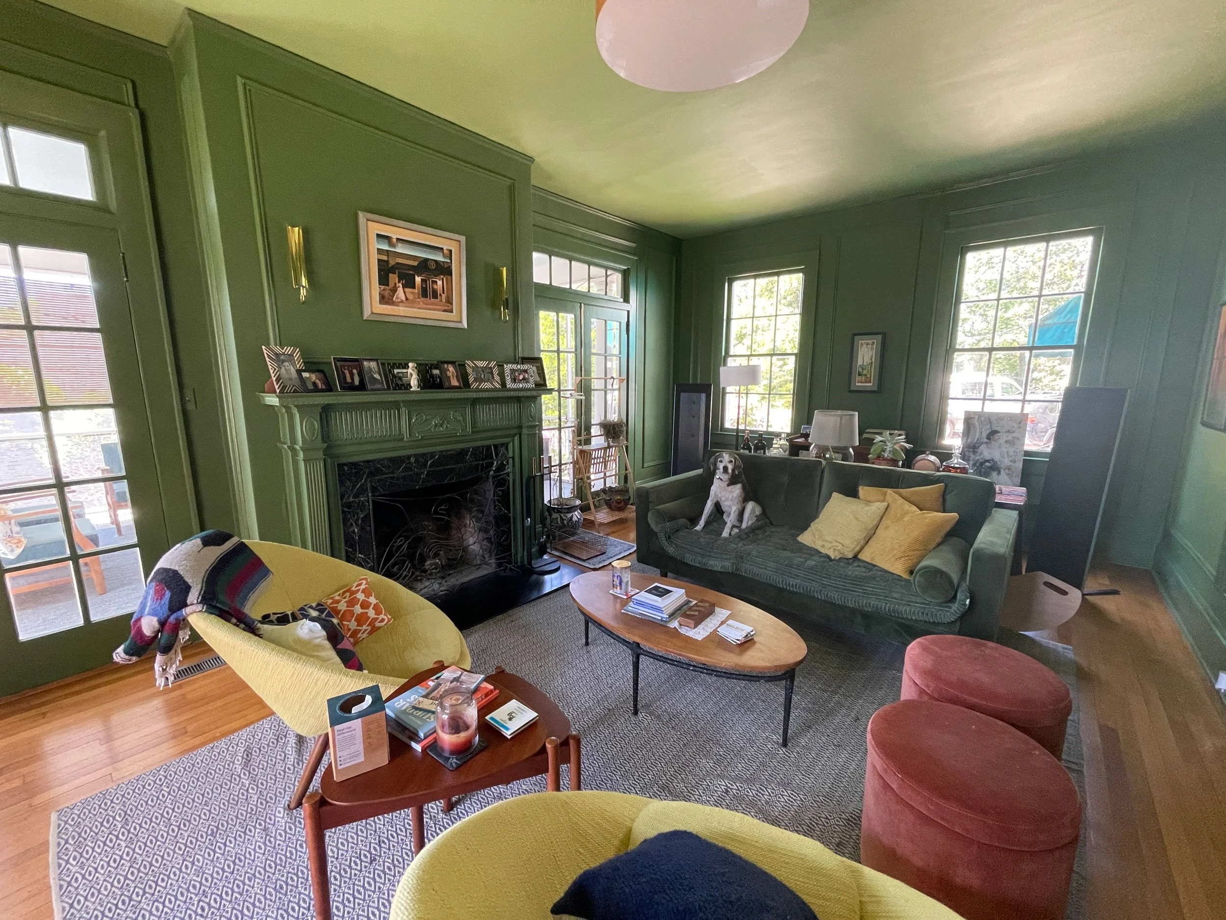 Living room with green walls and large windows, furnished with a yellow chair, a green sofa with yellow pillows, a wooden coffee table with books and a candle, and a red ottoman. A dog is sitting on the sofa. Decor includes framed photos on a mantel, a painting above the mantel, and various lamps and decorative items.