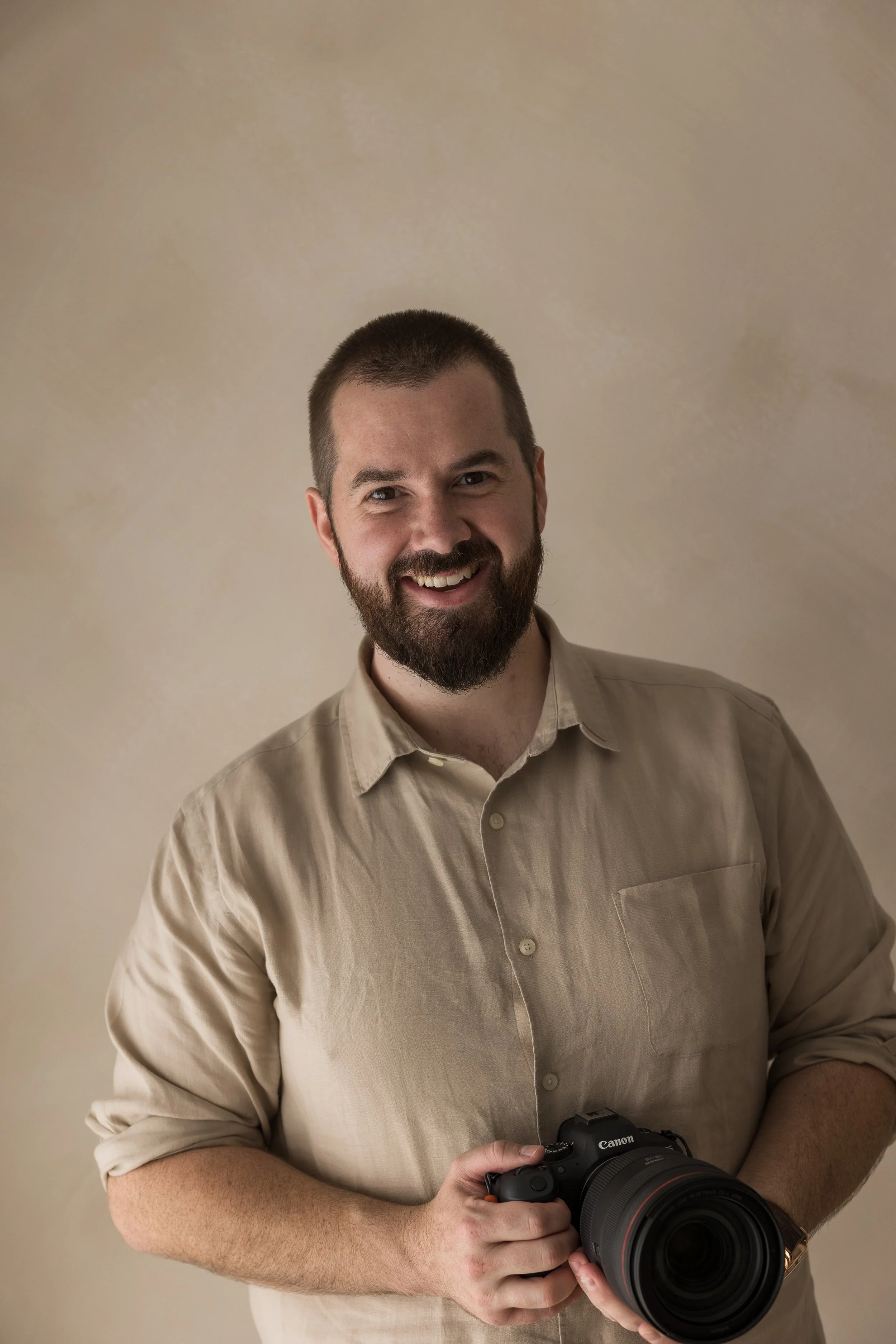 Smiling man with a beard holding a camera, wearing a beige button-up shirt, against a plain beige background.