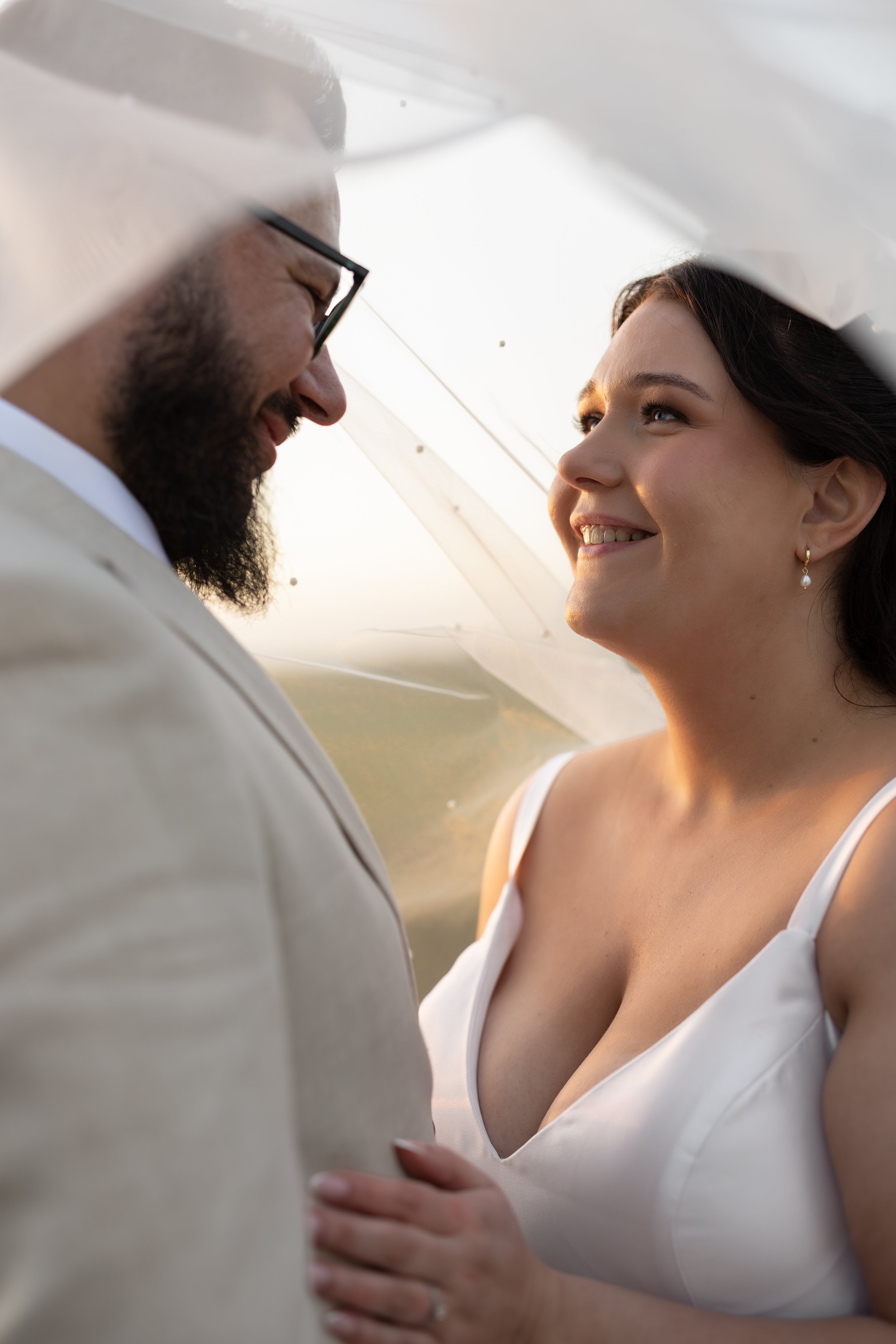Close-up wedding portrait of a bride and groom smiling at one another beneath her veil, with warm sunlight illuminating their faces.