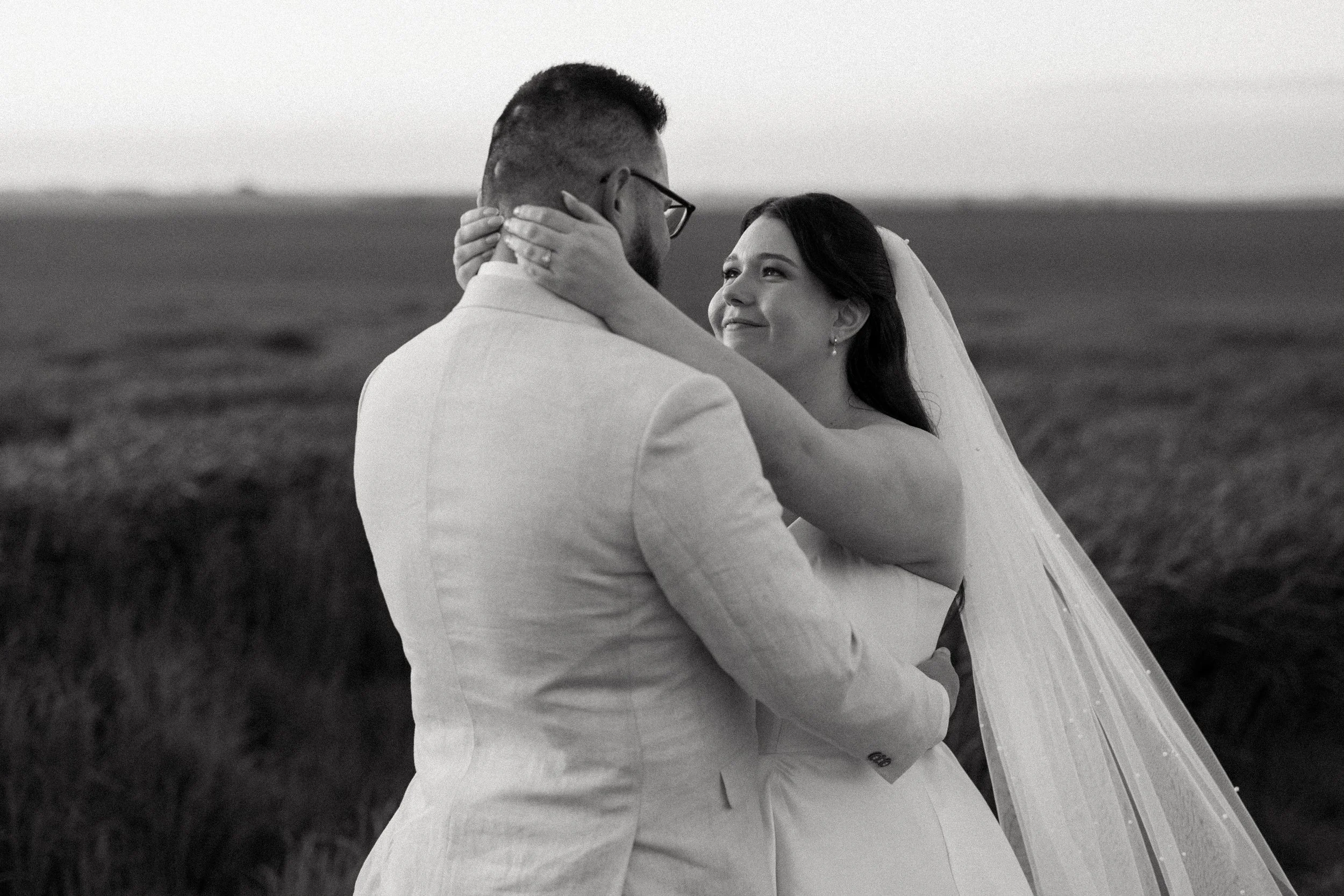 Black-and-white wedding photograph of a bride and groom embracing in an open field, with the bride looking up at her partner.