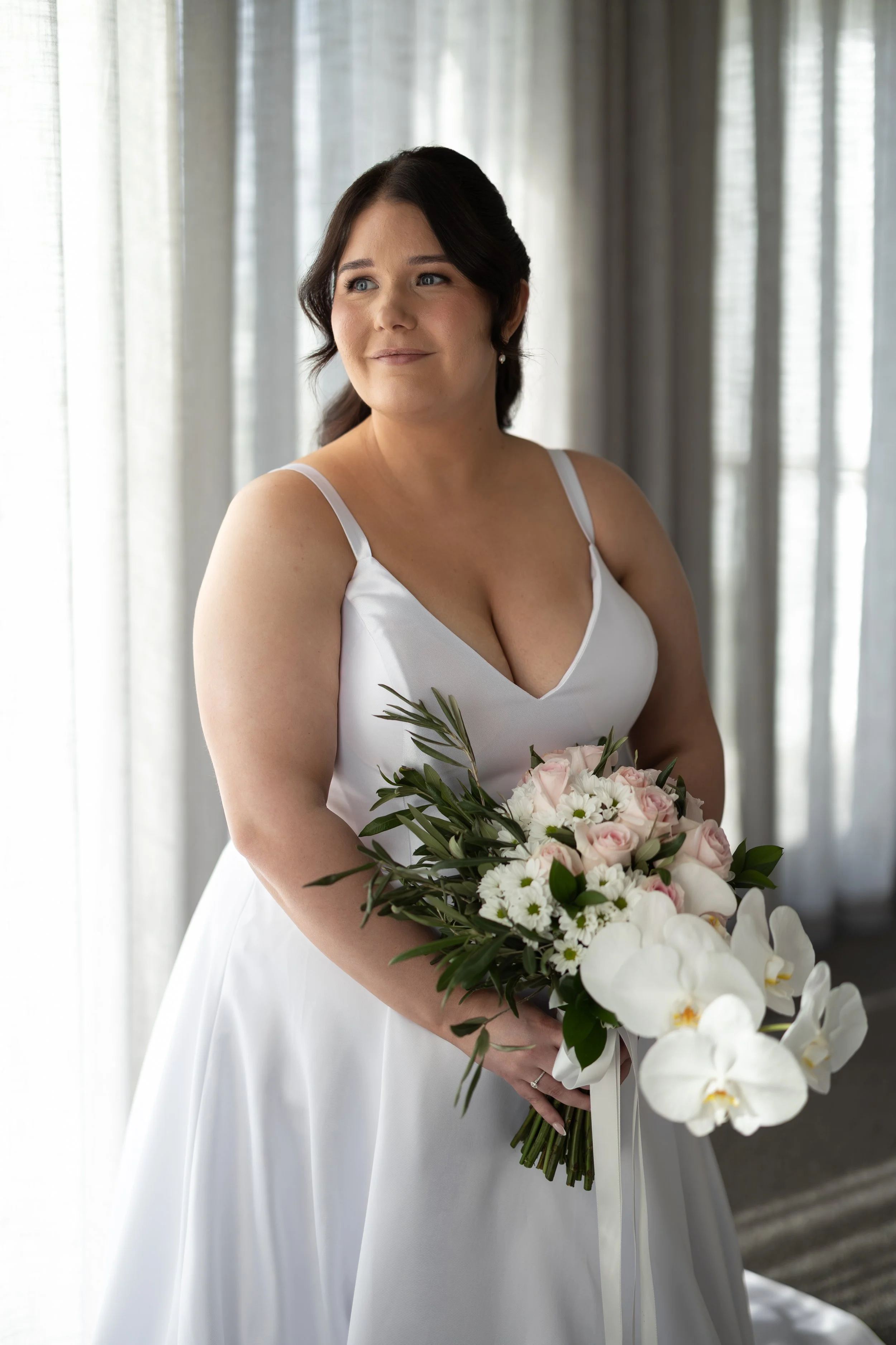 wedding-photography.jpg	Bride standing beside a window in a white wedding dress, holding a bouquet of pink roses and white orchids in soft natural light.