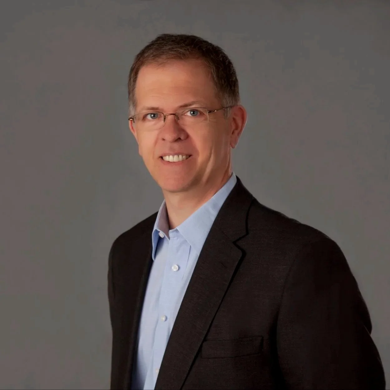 Portrait of a middle-aged man with short brown hair, glasses, wearing a dark suit jacket and light blue dress shirt, smiling against a plain gray background.